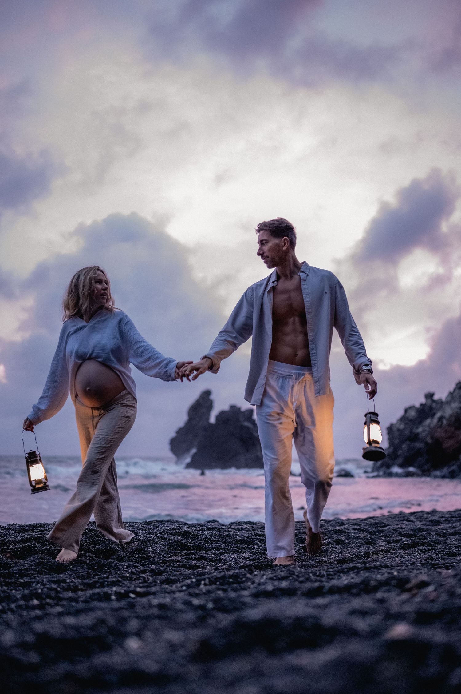 Couple holding lanterns walking on black sand beach at dusk, pregnant woman and man in light clothing.