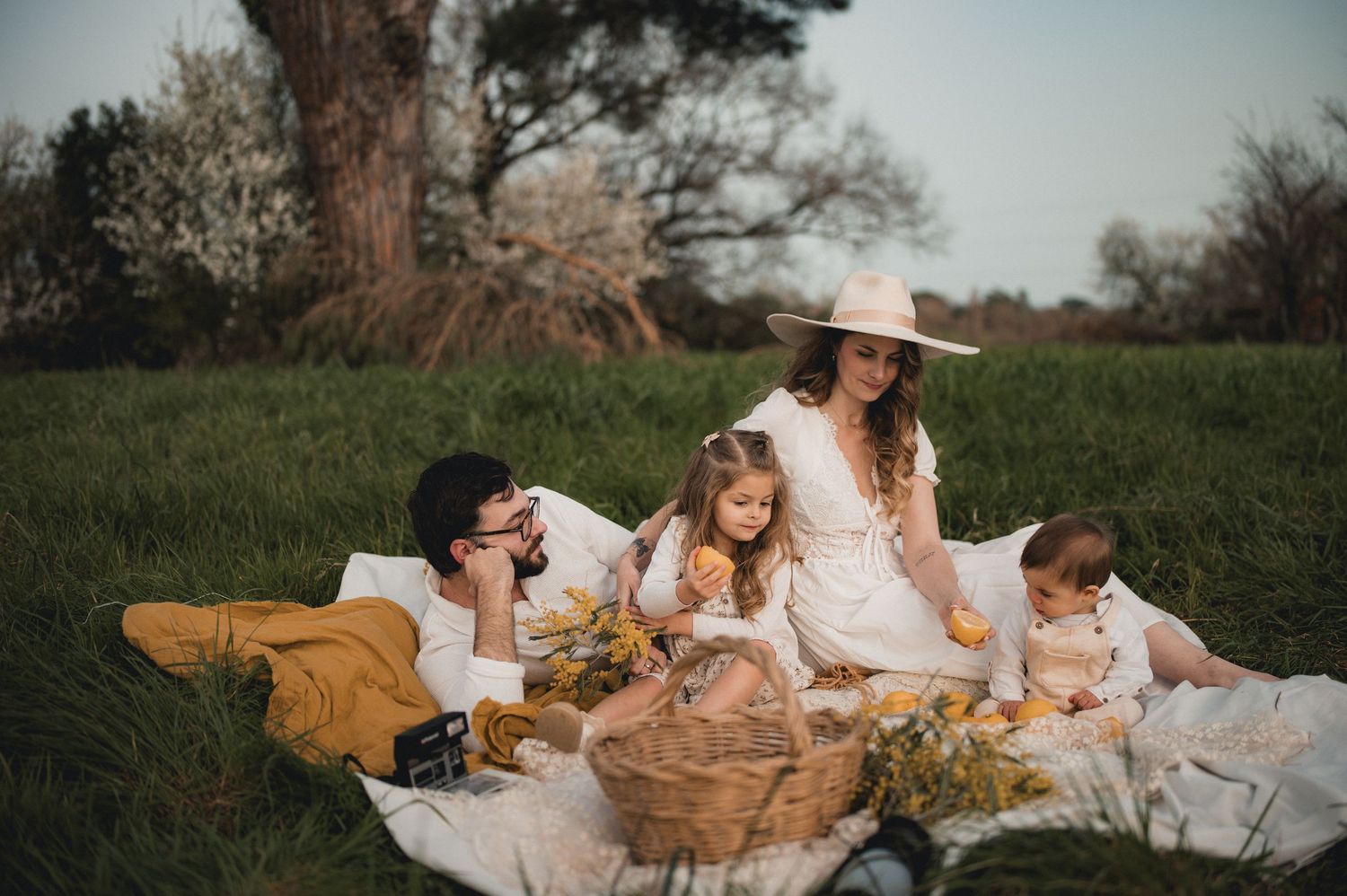 Family enjoying a picnic on a white blanket in a green meadow with a wicker basket of flowers.