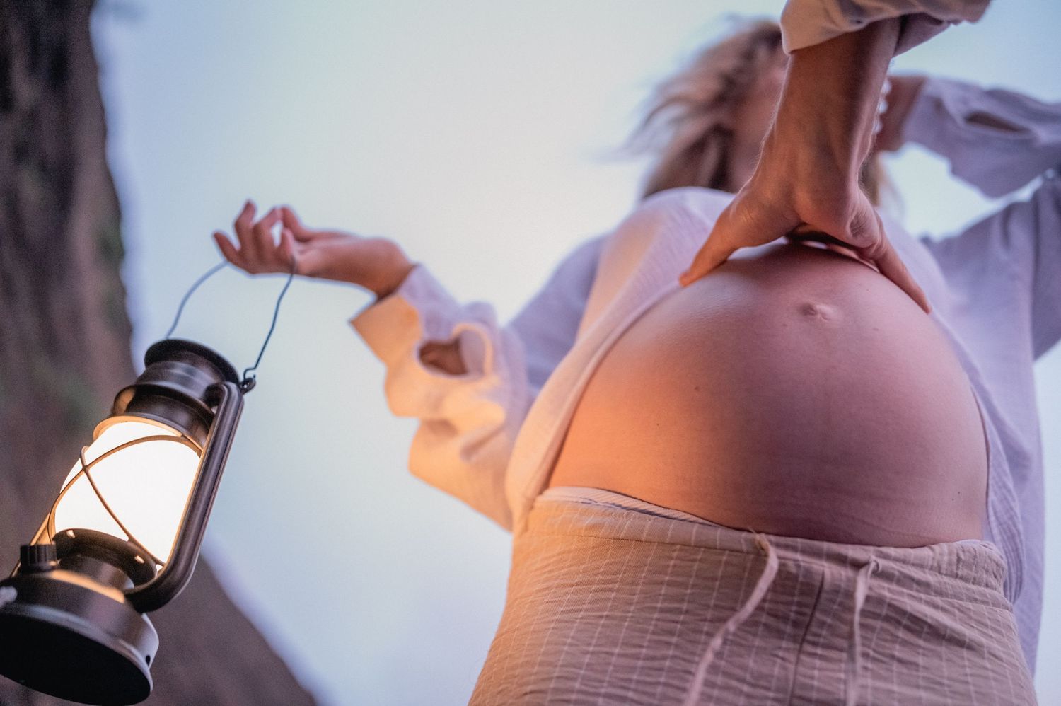 Close-up of pregnant woman holding vintage lantern, showing bare baby bump in white shirt.