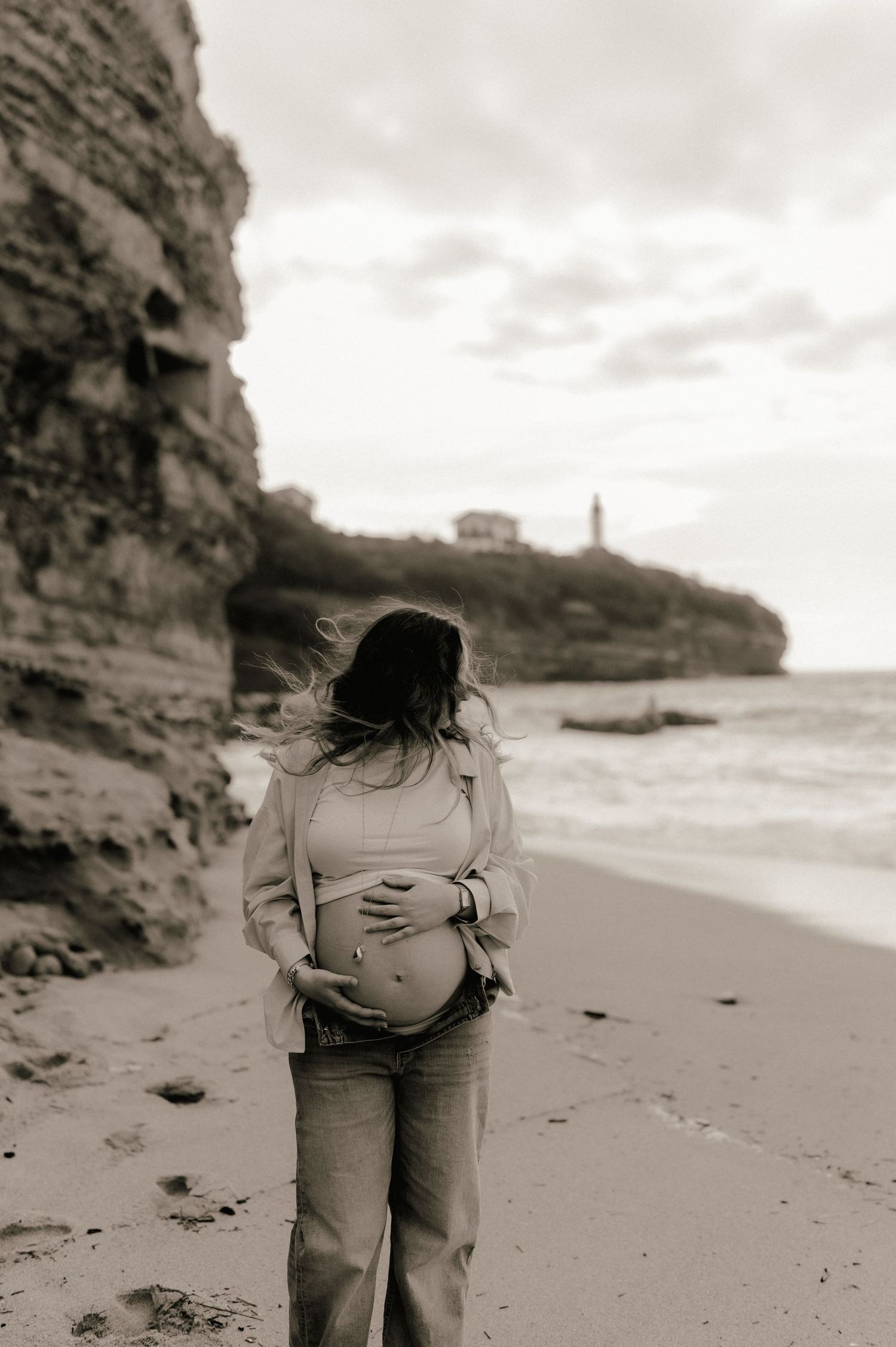 Sepia photo of a pregnant woman cradling her belly on a windy beach with rocky cliffs behind.