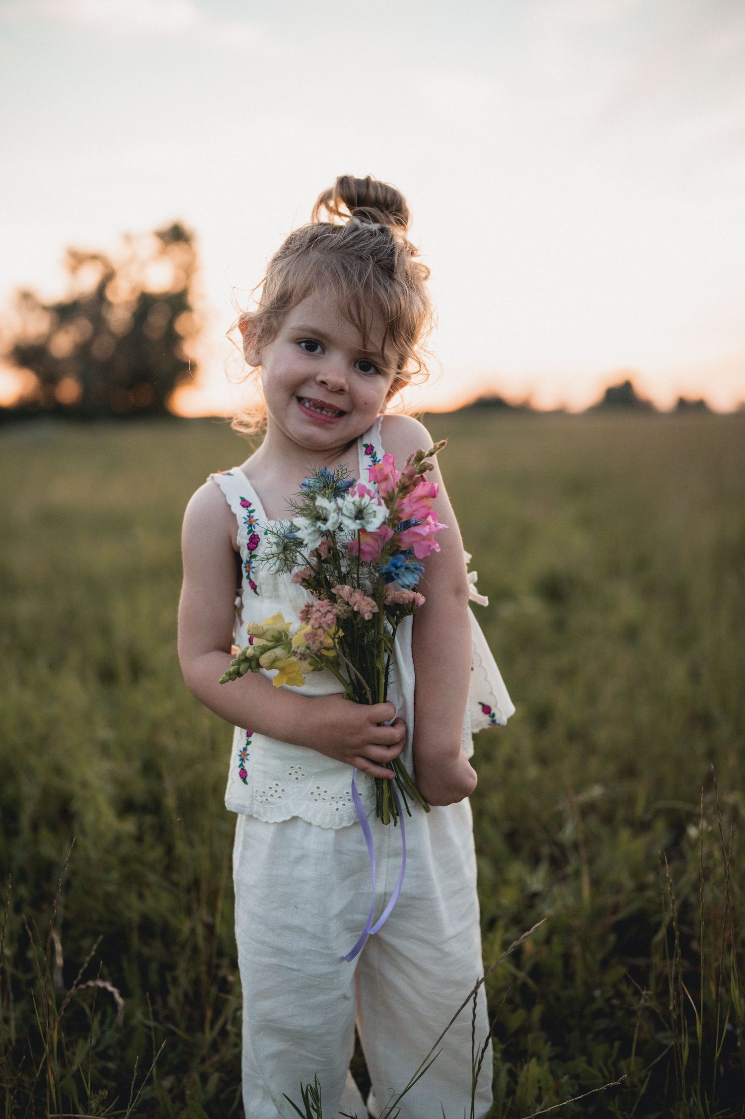 A smiling little girl in white holds a bouquet of wildflowers in a golden sunset field.