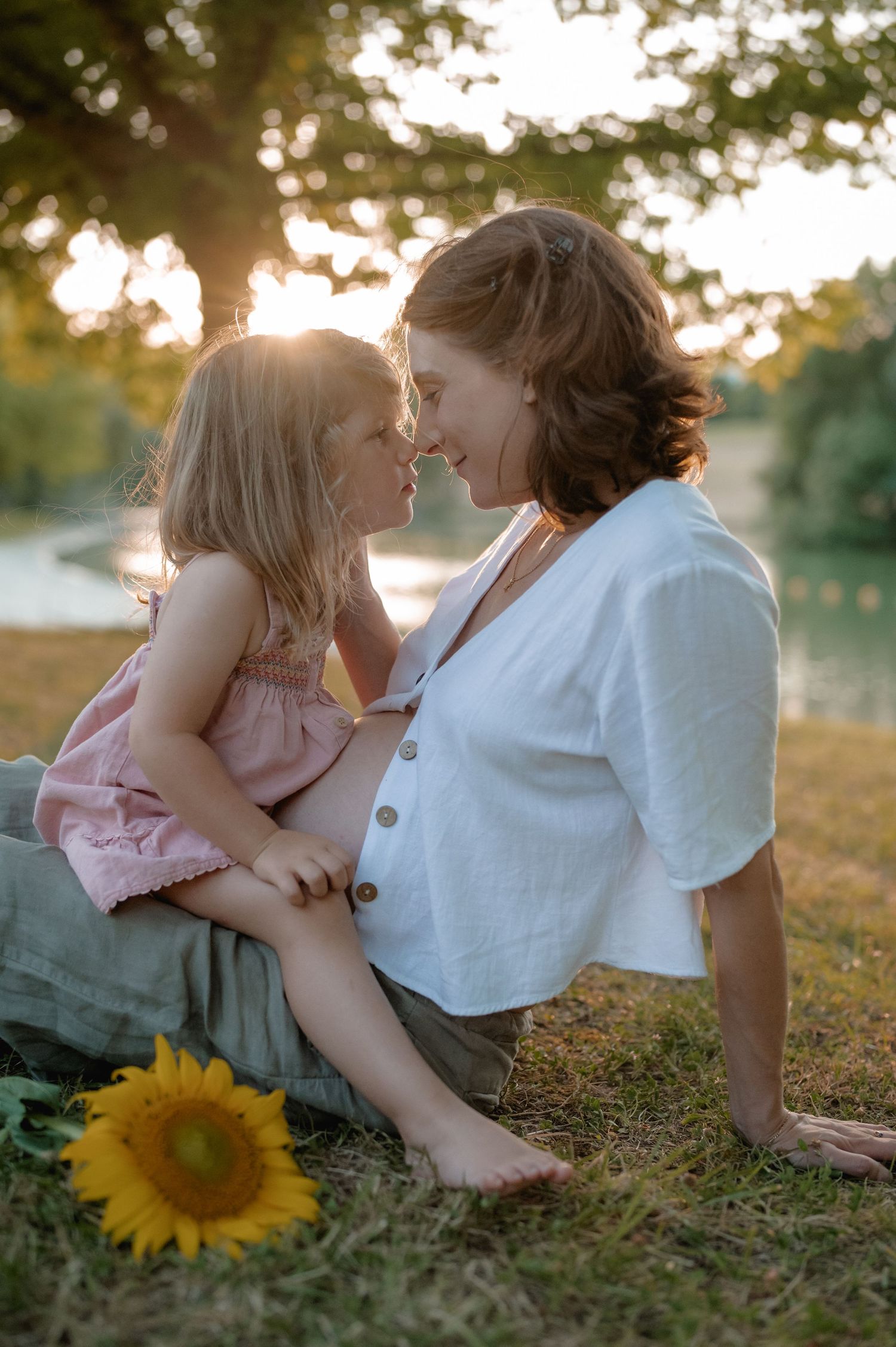 Mother and daughter touching noses tenderly in golden sunset light near sunflower by riverside.