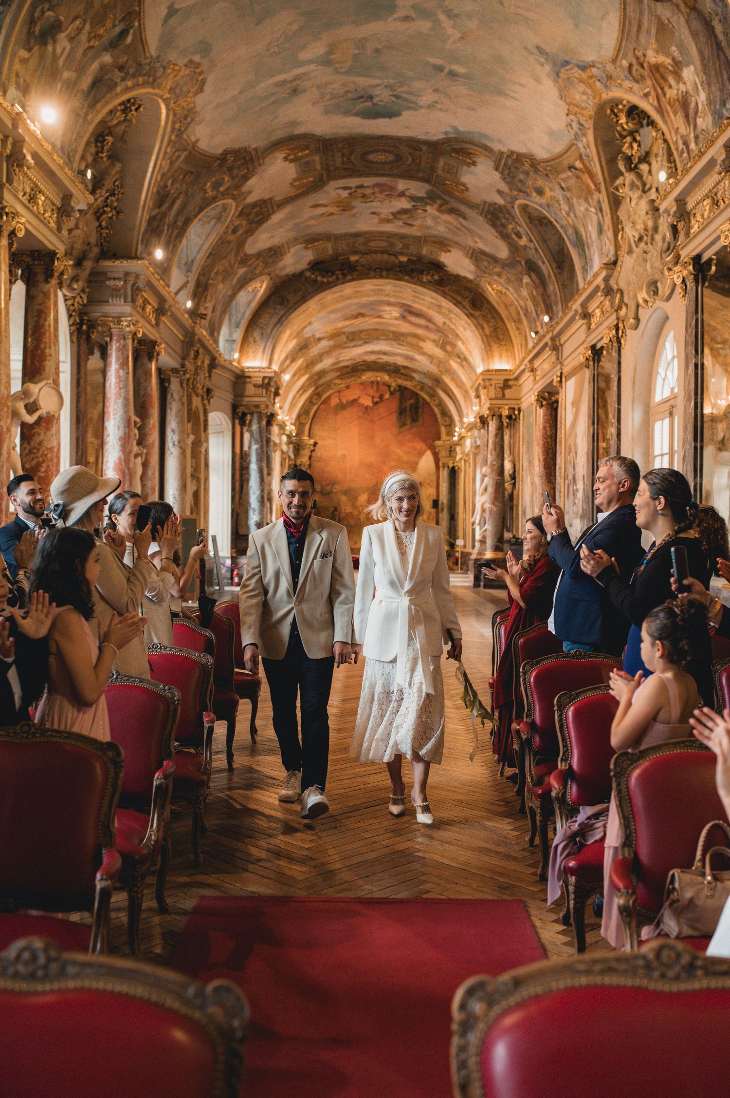 Newlyweds walking down grand baroque hall aisle surrounded by applauding guests in red velvet chairs.