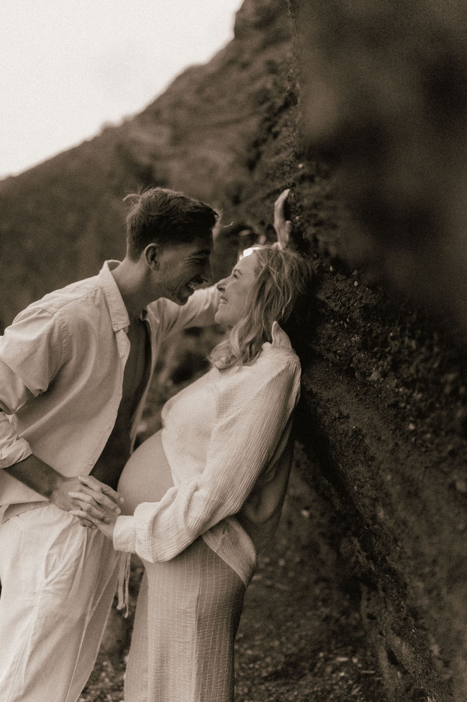 Couple sharing a tender moment leaning against a mossy rock face in sepia-toned maternity photo.