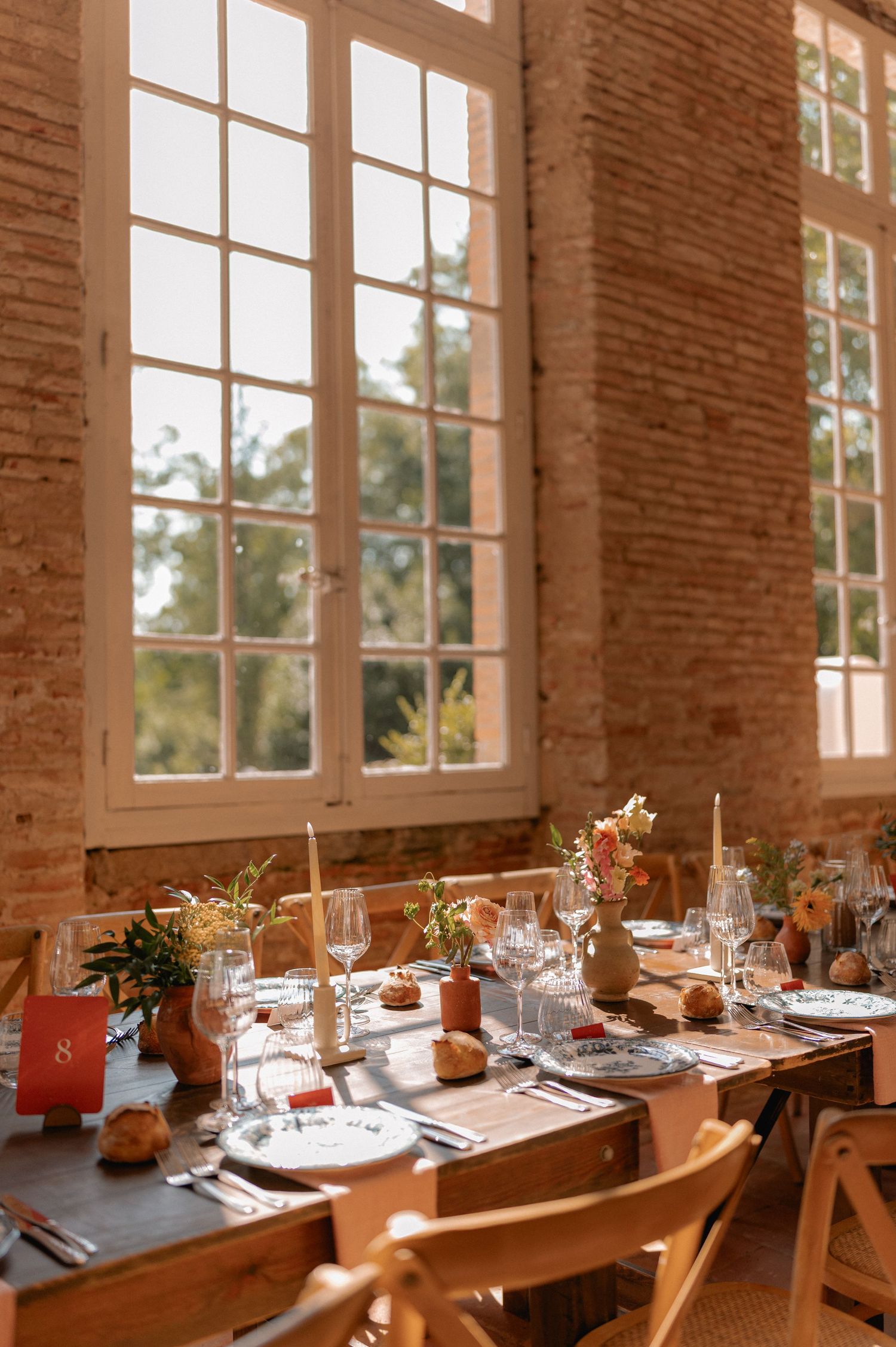Elegant rustic wedding reception table set with glassware, candles, and floral centerpieces in a sunlit brick venue.