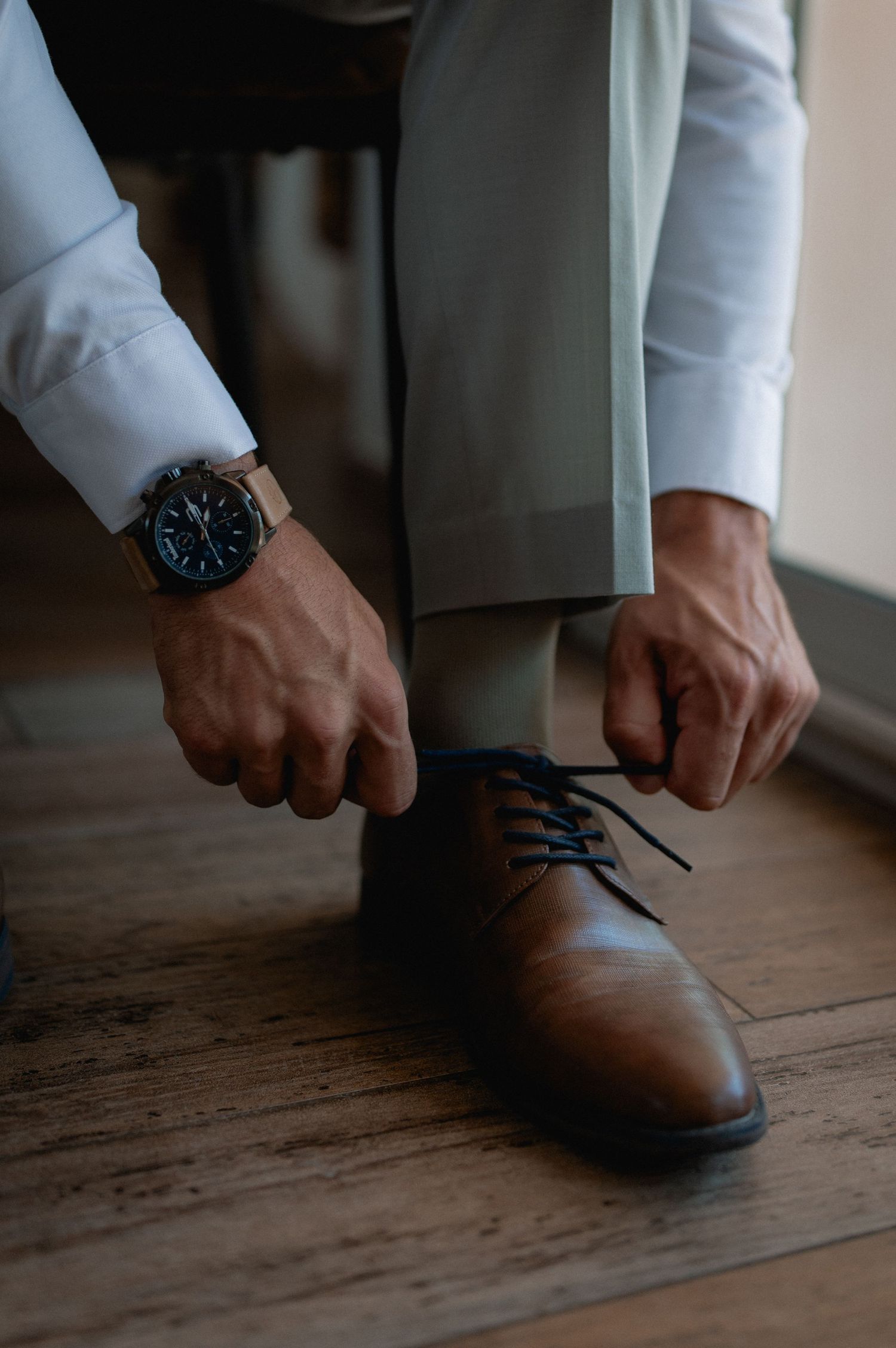 Man in white shirt and gray trousers tying brown leather Oxford shoes with navy laces, wearing a black analog watch.