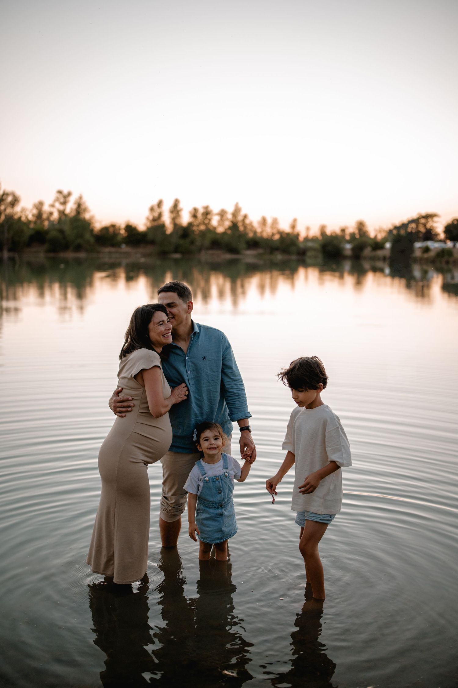 Happy family with pregnant mother, father, and two children standing in calm lake at sunset.