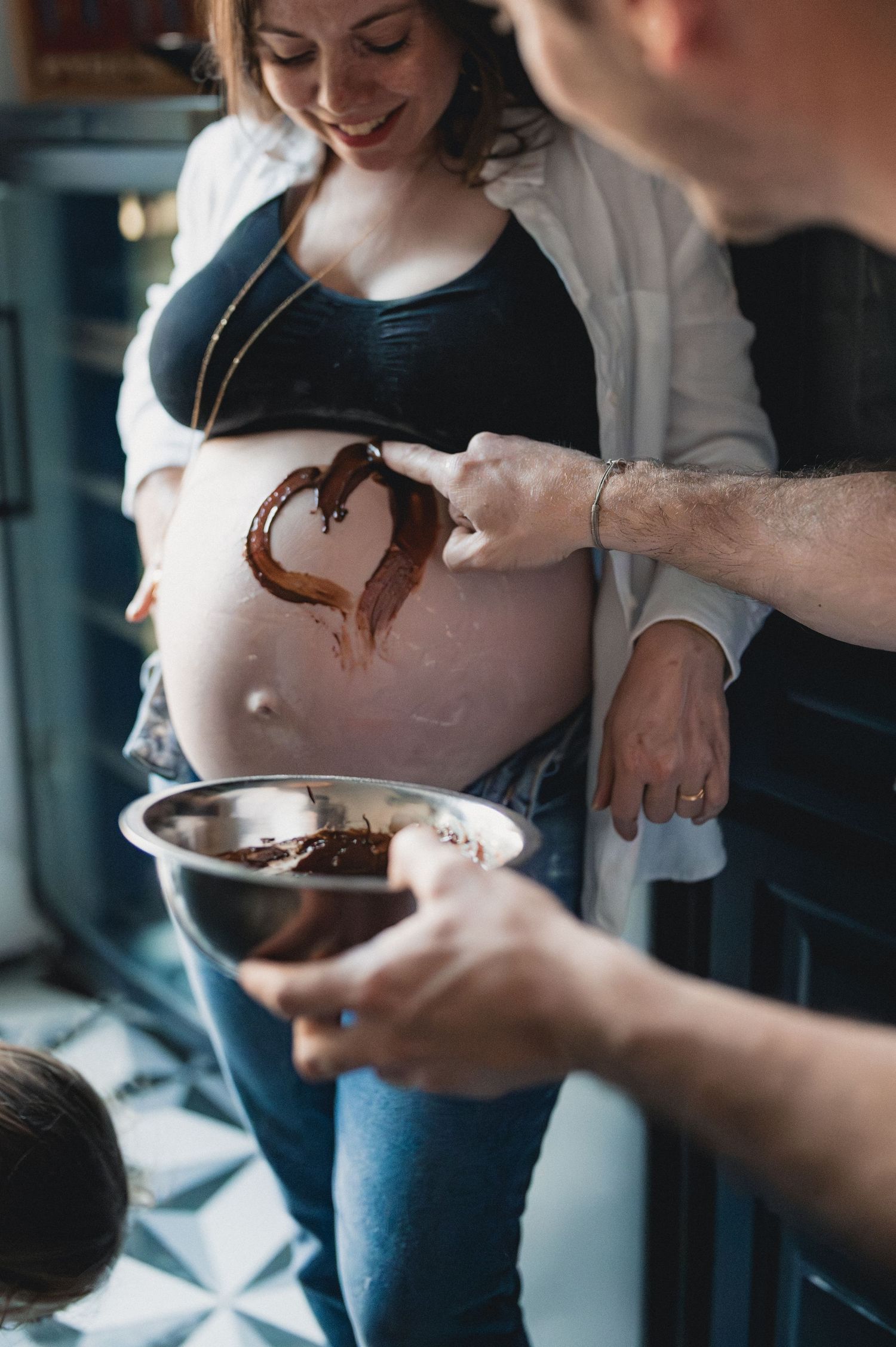 Pregnant woman with chocolate heart painted on baby bump by partner in kitchen.