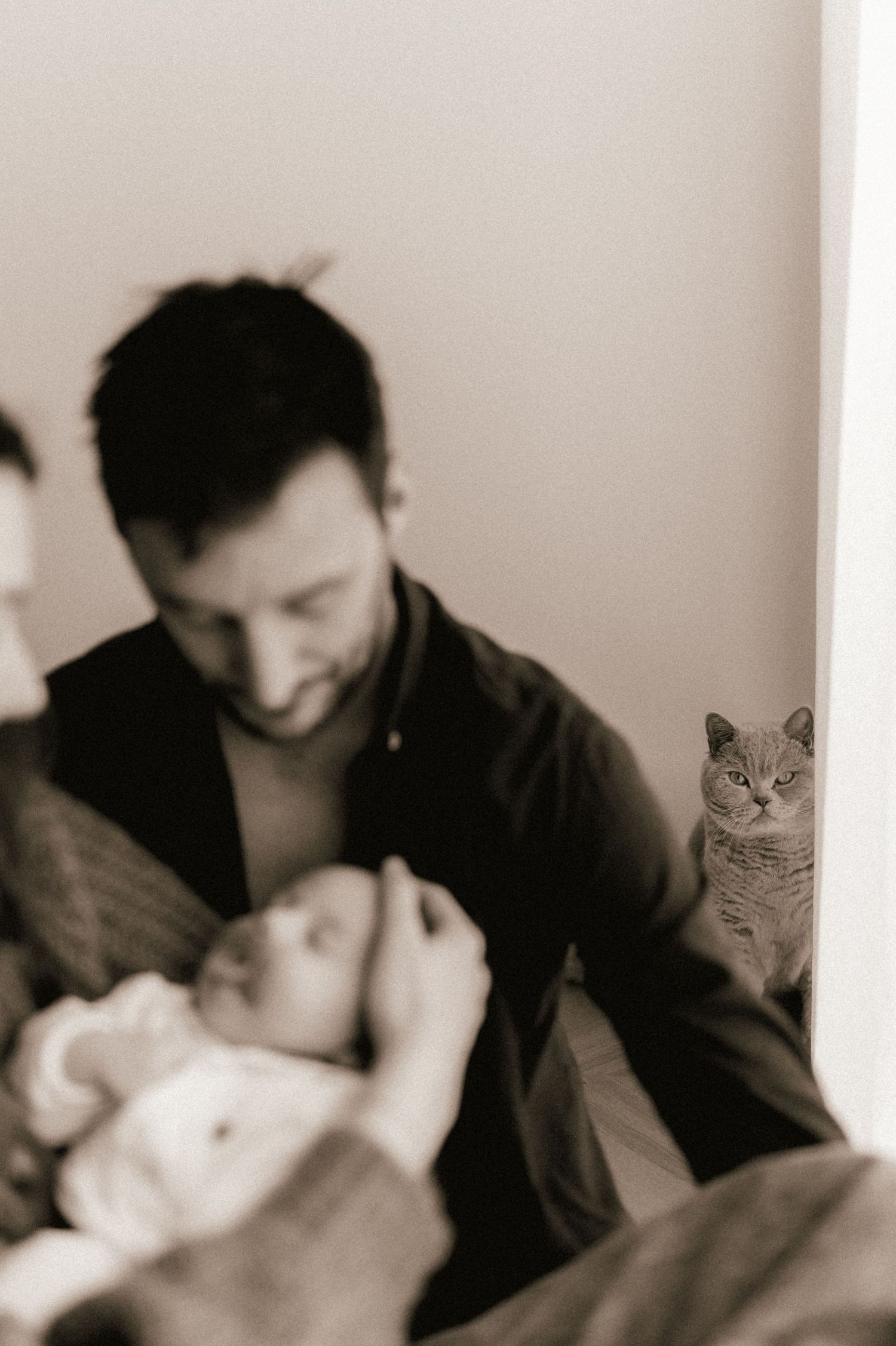 Father cradling newborn baby while a curious cat watches in the background, black and white photo.