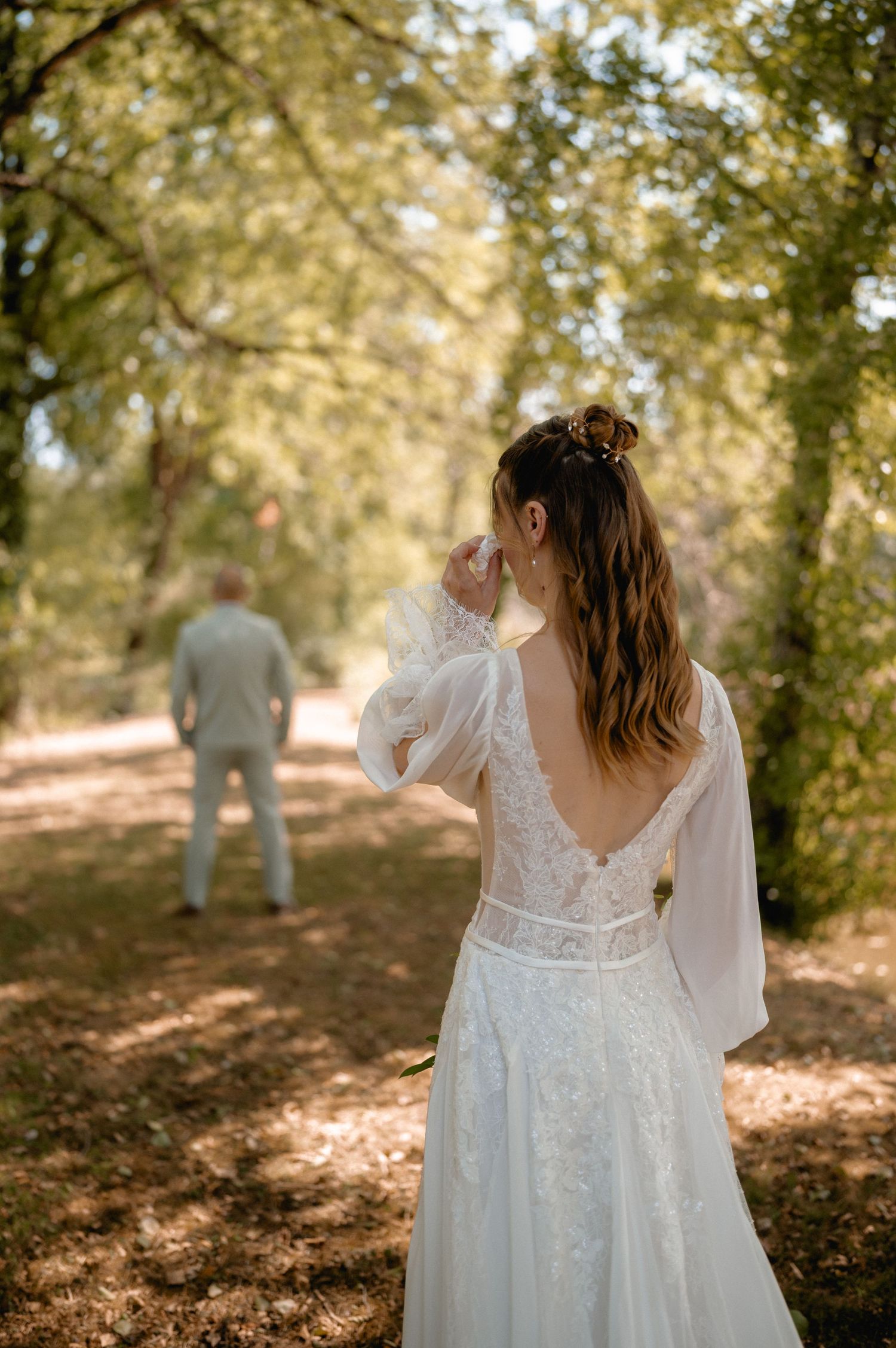 Bride in white lace gown with open back stands on forest path, groom visible in distance.