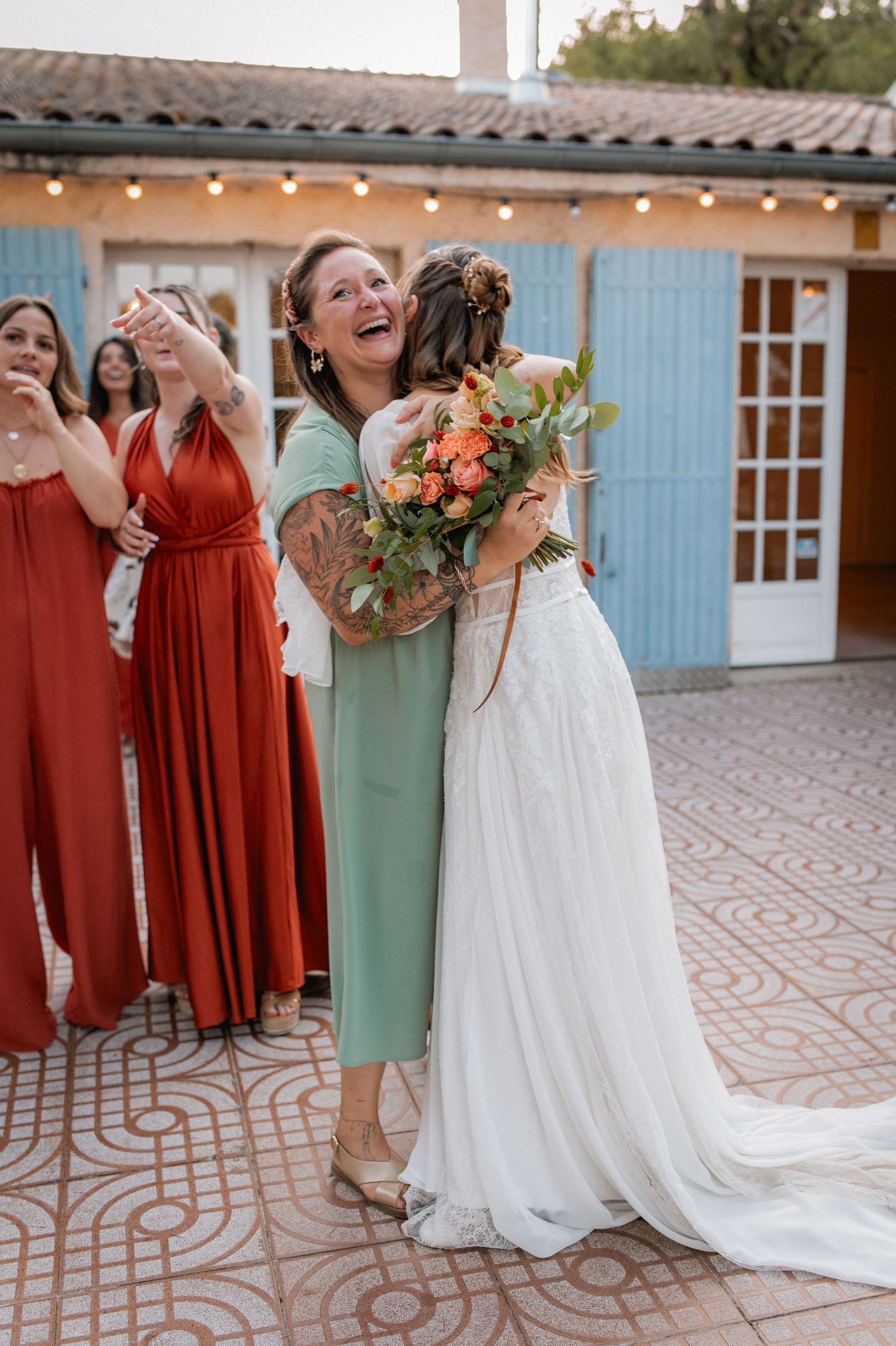 Bride in white gown hugging guest in green dress, laughing, with bridesmaids in red dresses at outdoor wedding venue.