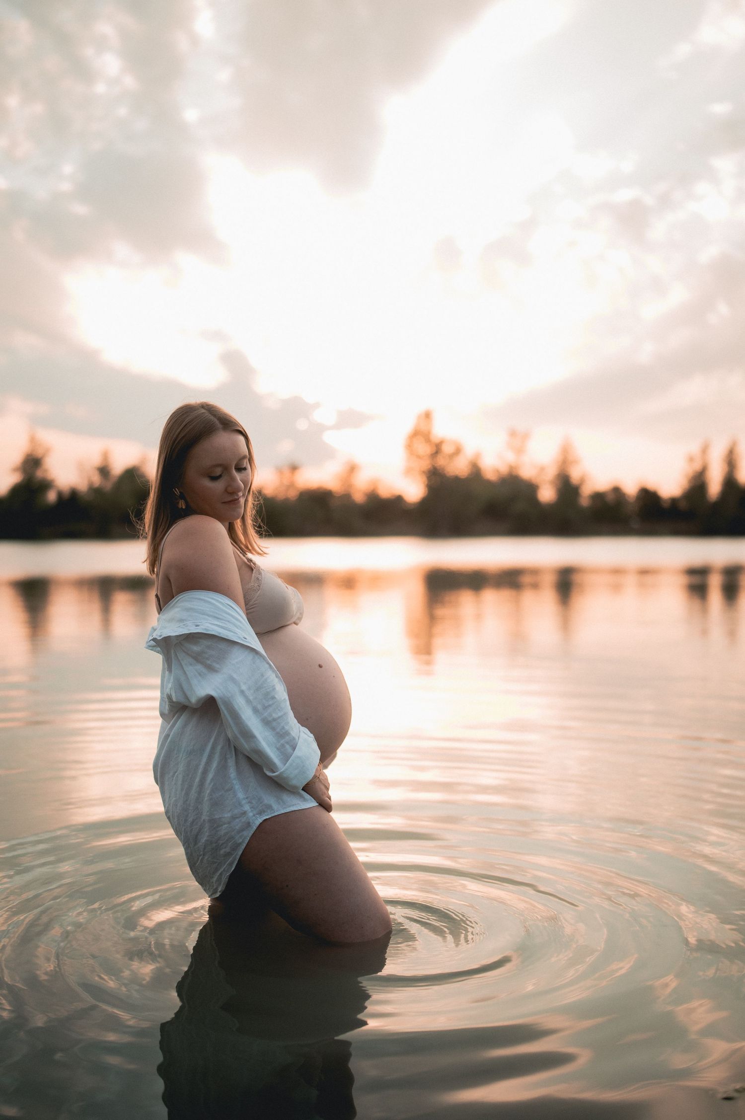 Pregnant woman standing in a calm lake at sunset, cradling her belly in an open white shirt.