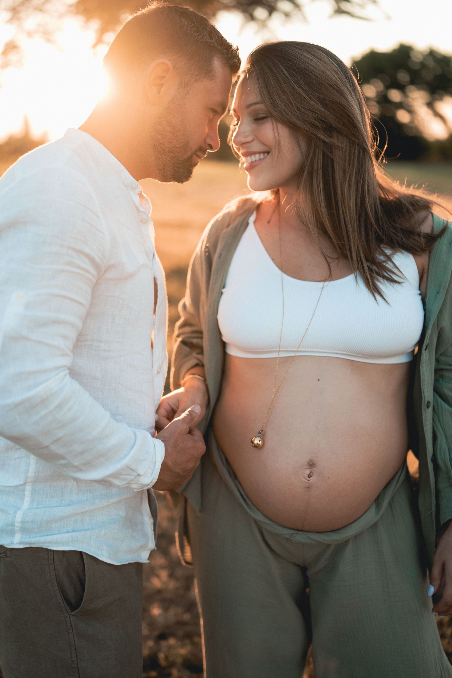 Smiling pregnant woman in white crop top holds hands with man at golden hour sunset outdoors.