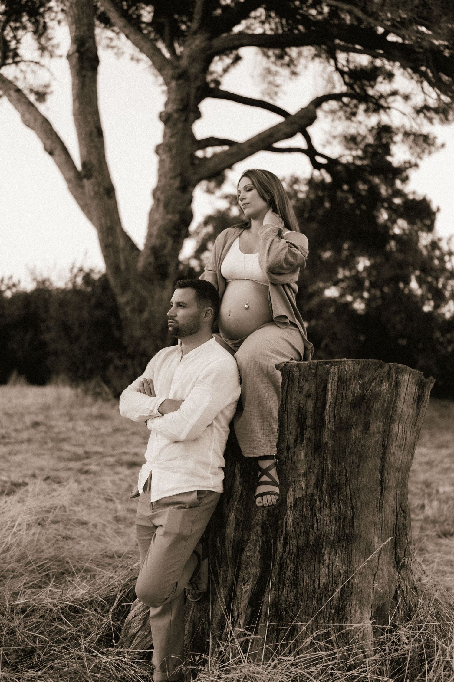 Sepia maternity photo of a couple near a large tree stump in a natural outdoor setting.