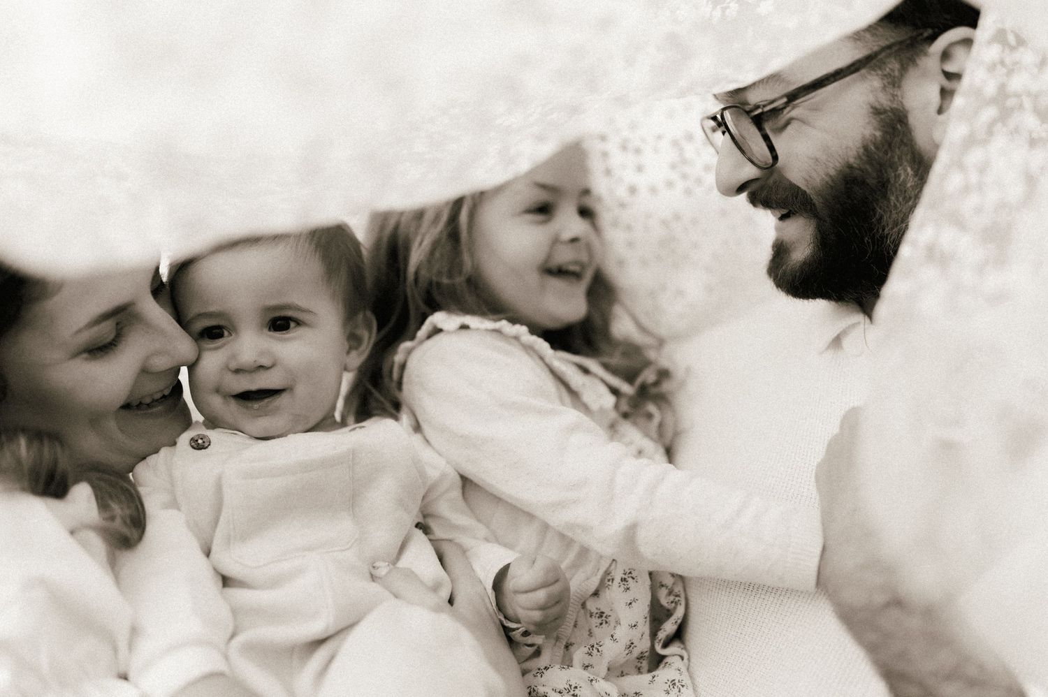 Black and white photo of a happy family with two young children snuggled together under a white blanket.