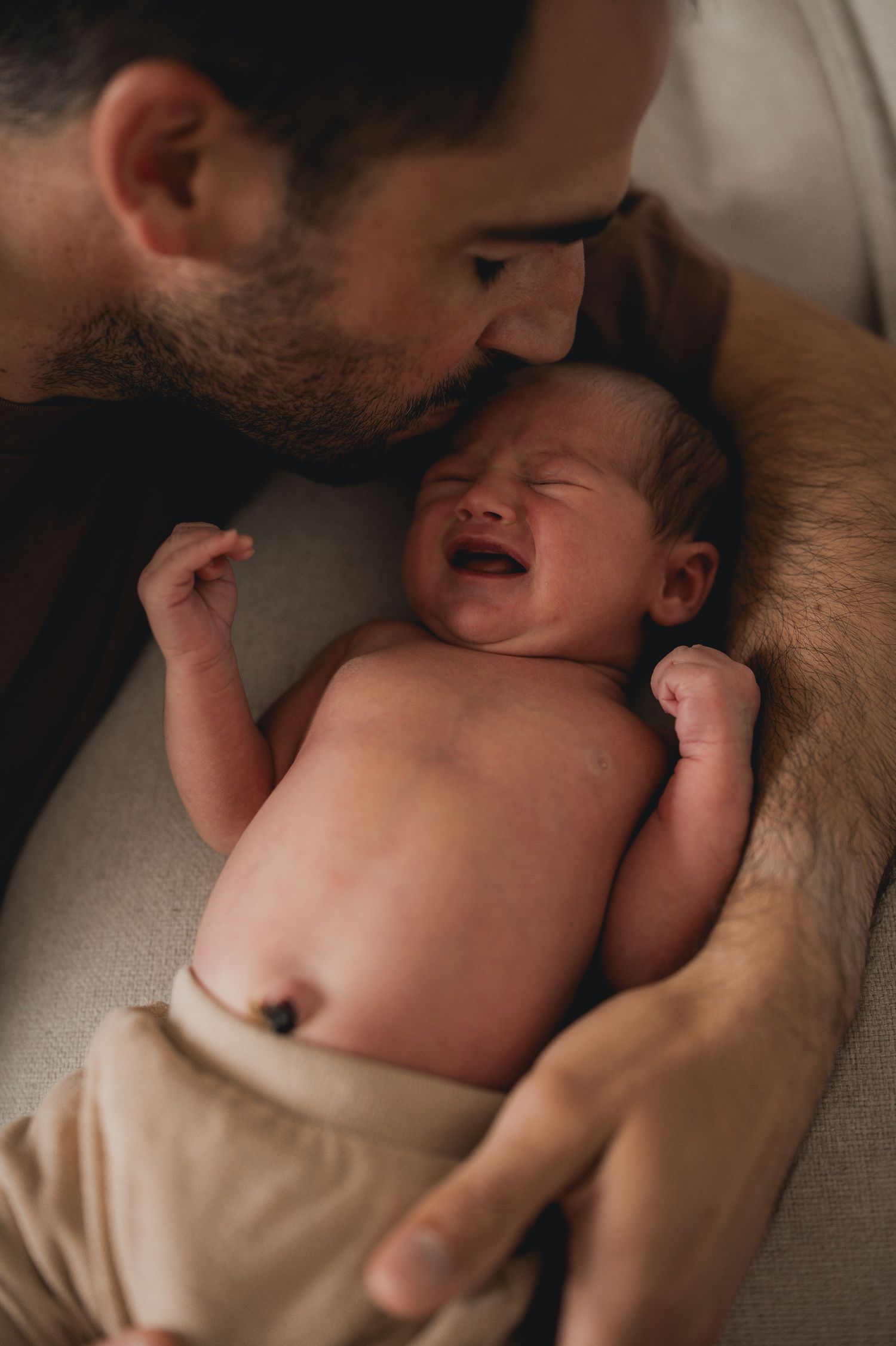 Father gently kissing his crying newborn baby lying on his chest.