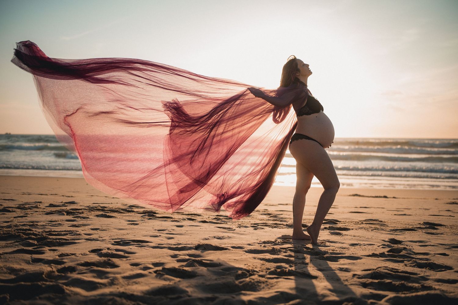 Pregnant woman in bikini poses on beach at sunset with flowing red fabric billowing in the wind.