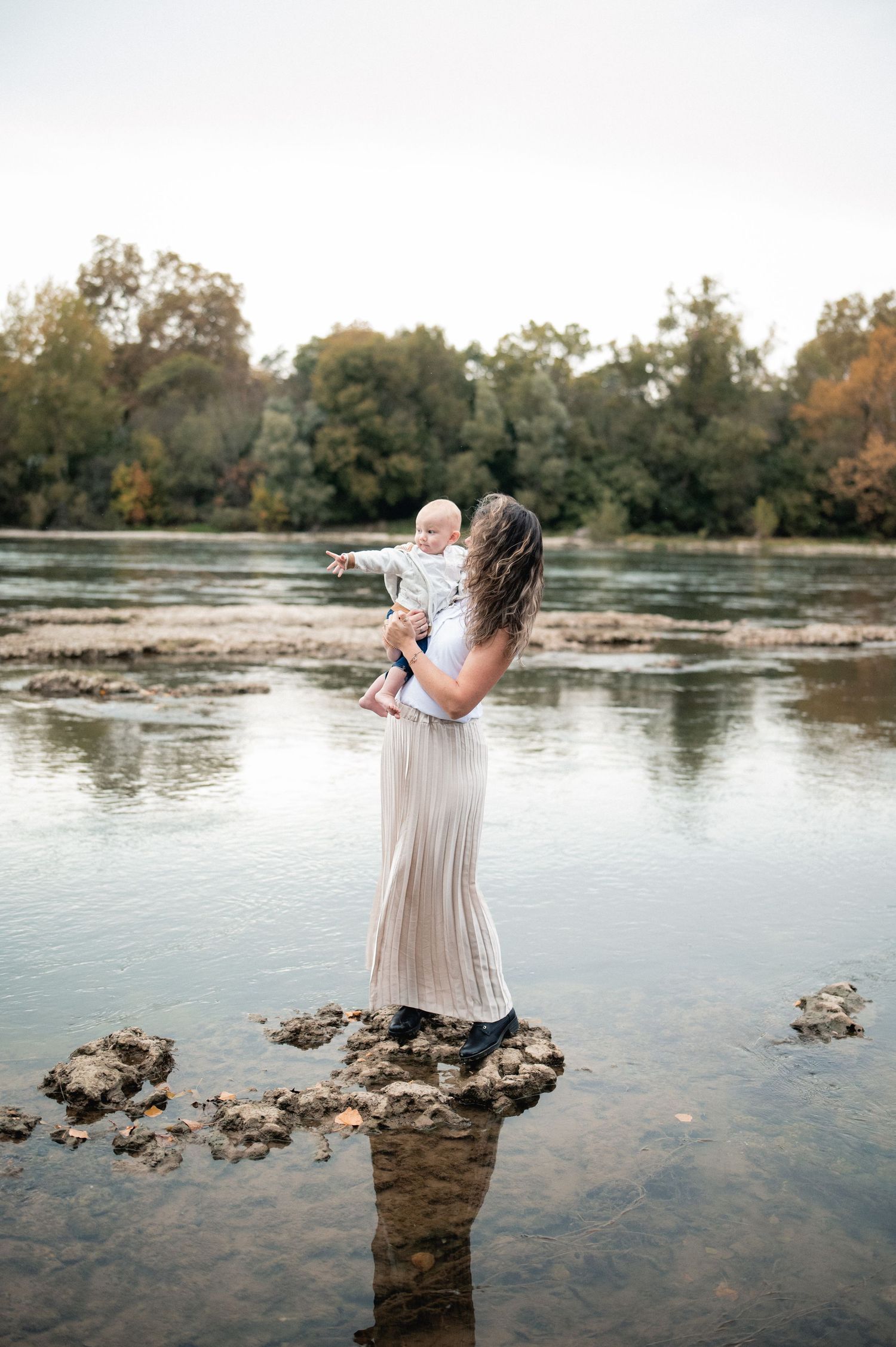 Mother in flowing beige dress holds baby while standing on rocks beside a calm river.