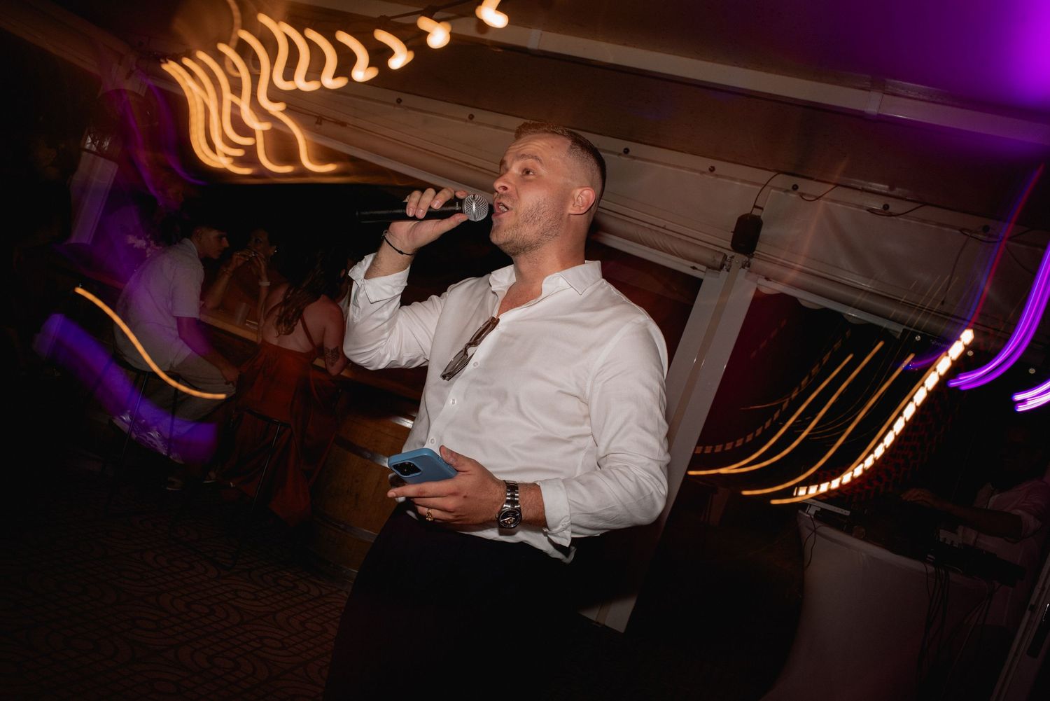 Man in white shirt singing karaoke with microphone at a vibrant nightclub event with colorful lights.