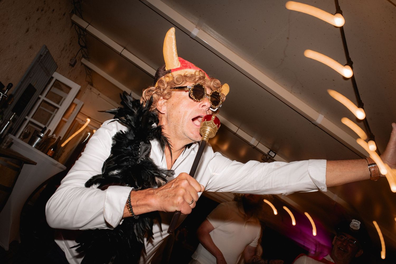 Person in costume with devil horns, curly wig, sunglasses, and feather boa dancing at a party.