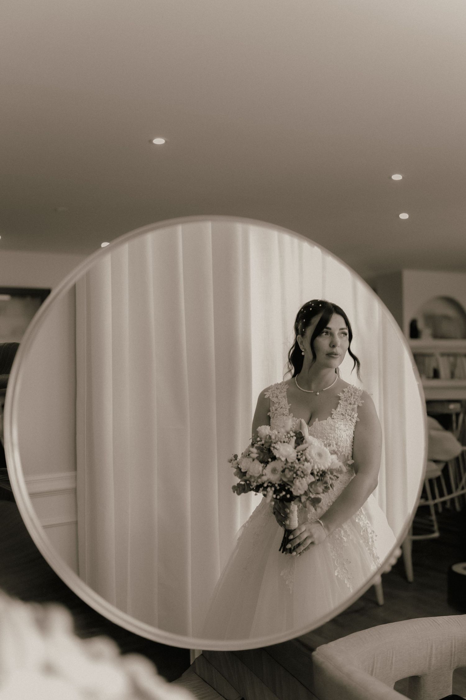 Black and white mirror reflection of a bride in lace wedding dress holding a floral bouquet, standing by sheer curtains.