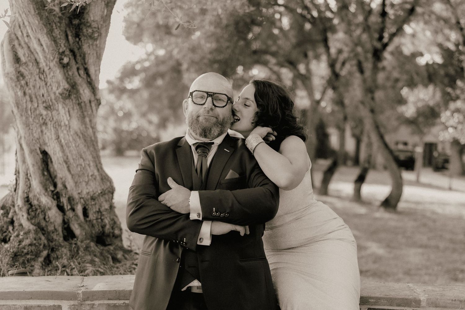 Bald groom in suit receives kiss on cheek from bride in black and white photo.