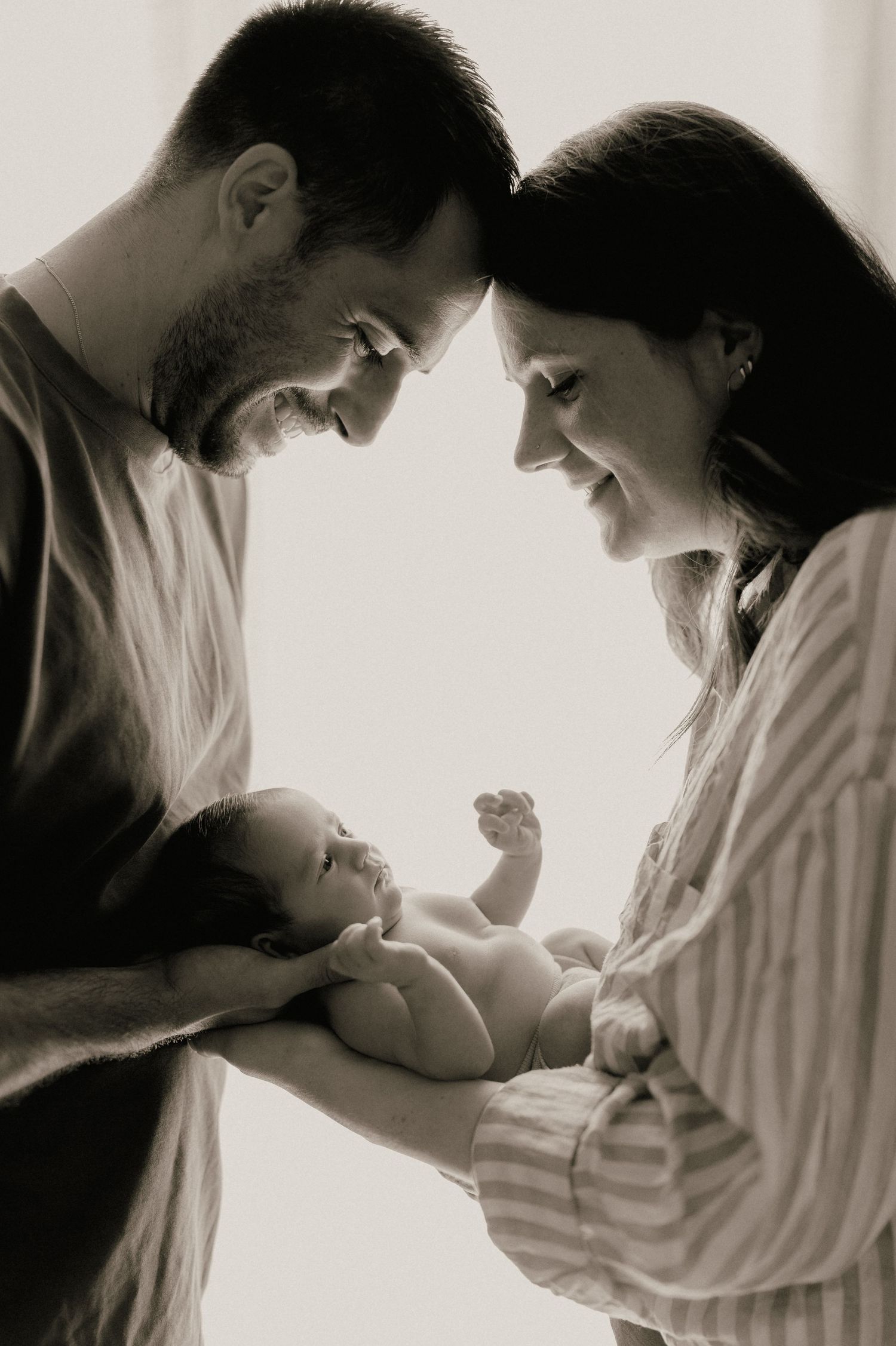 Black and white photo of new parents smiling down at their newborn baby held between them.
