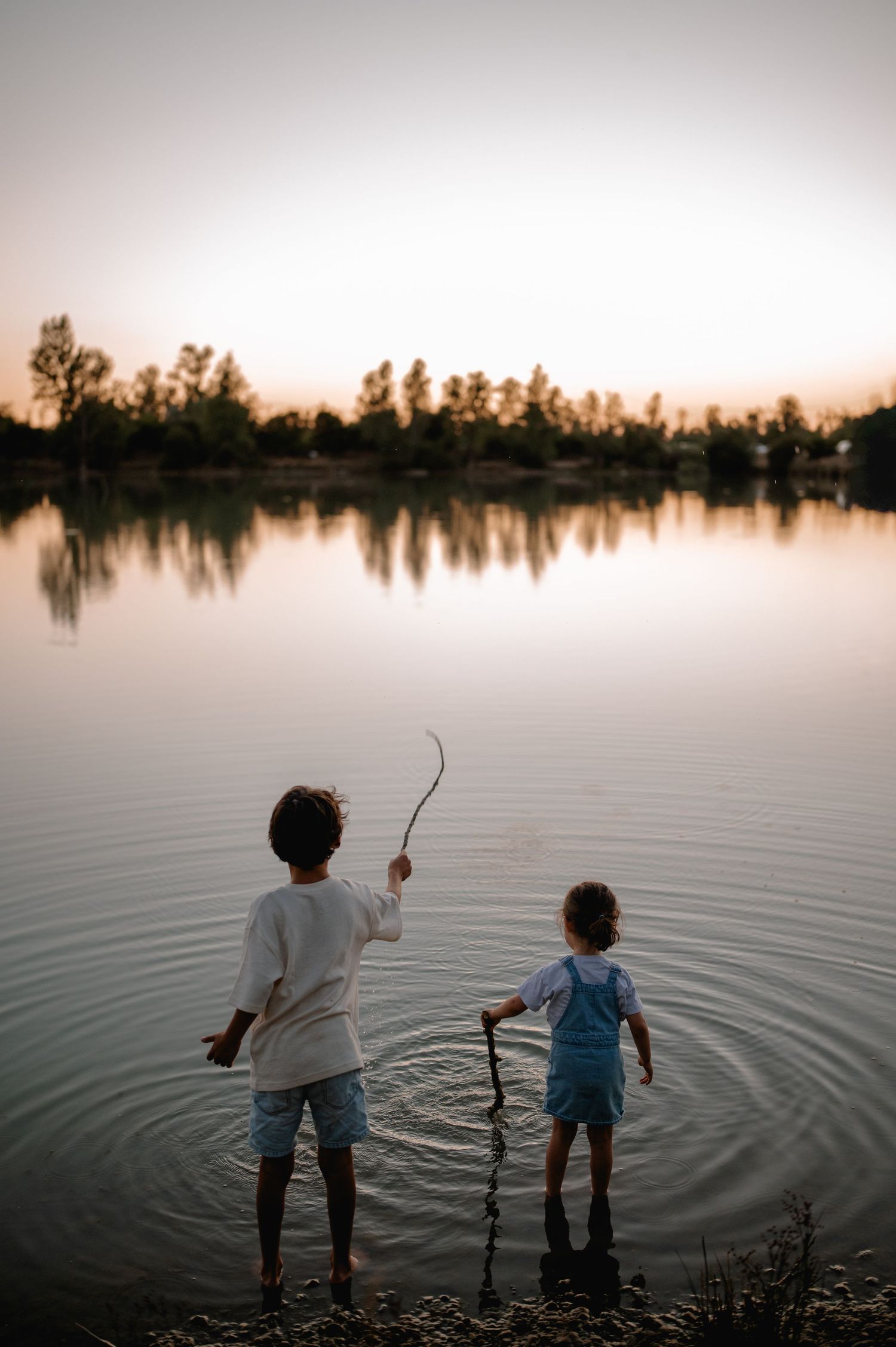 Two young boys stand at the edge of a calm lake at sunset, playing with sticks in the water.