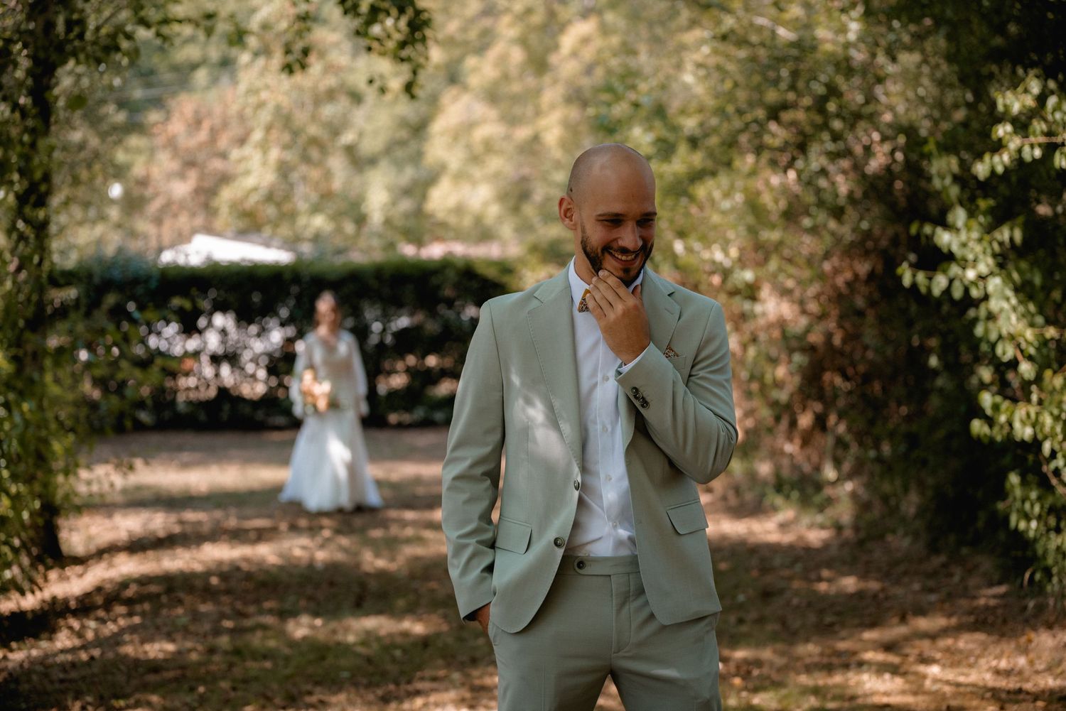 Groom in sage green suit smiling emotionally during first look moment in a lush garden setting.