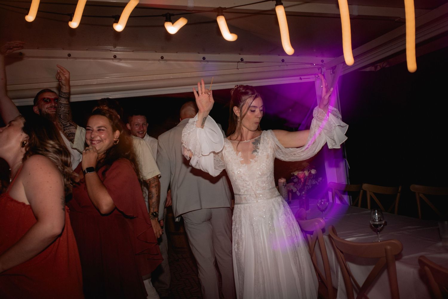 Bride in white wedding gown dancing joyfully with hands raised at an outdoor reception with guests in burgundy attire.