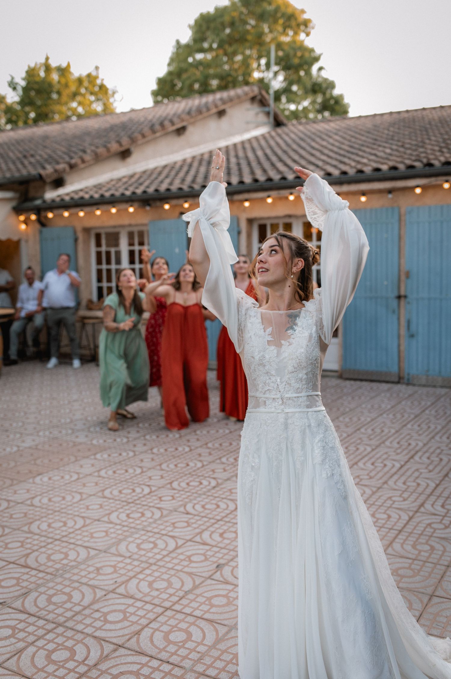 Bride in white lace gown tossing bouquet to guests in colorful dresses at outdoor wedding venue.