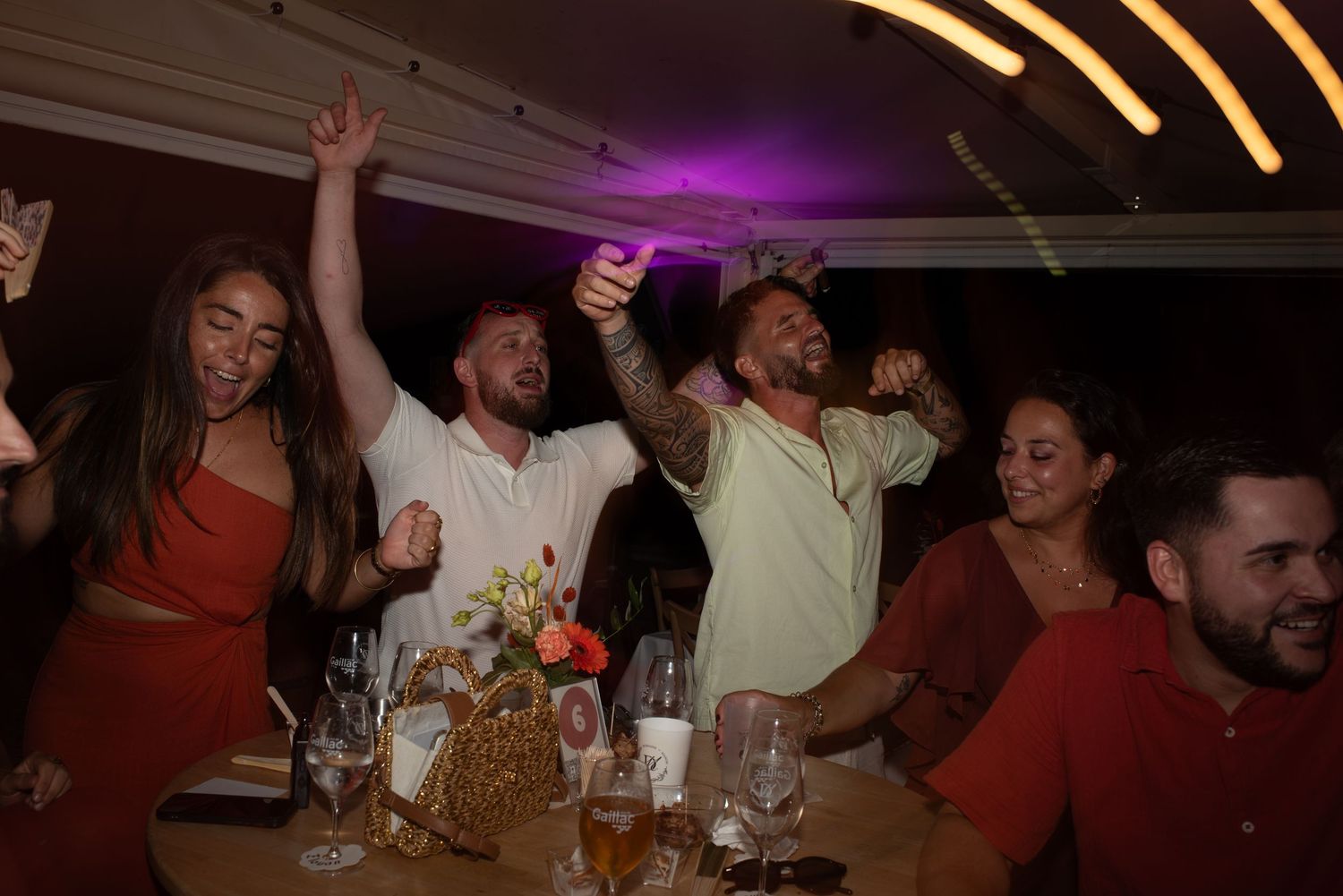 Group of friends celebrating at a dinner party, raising arms and cheering with drinks at a festive indoor event.