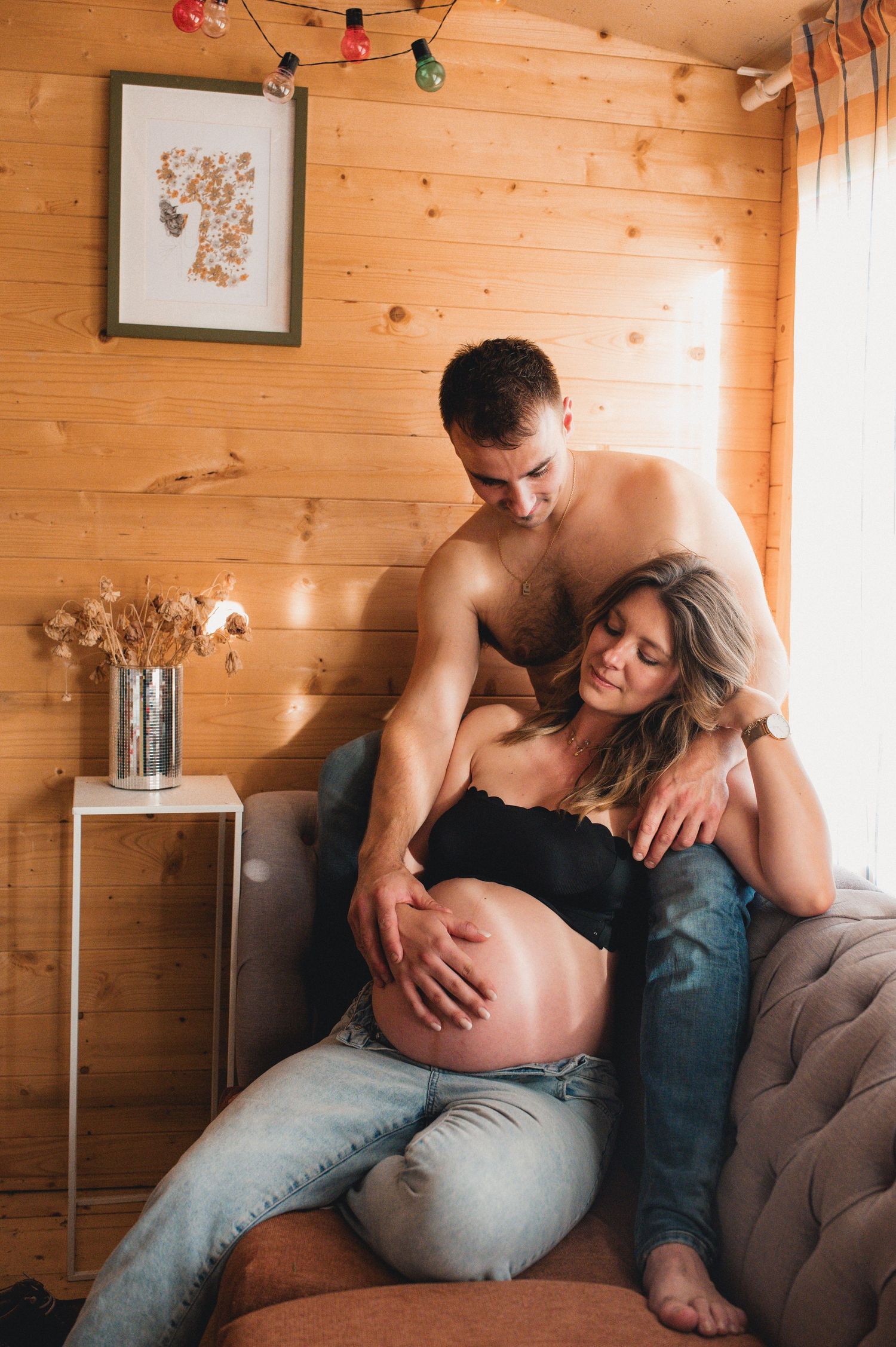 Couple in cozy wooden cabin, shirtless man embracing pregnant woman in black top and jeans.
