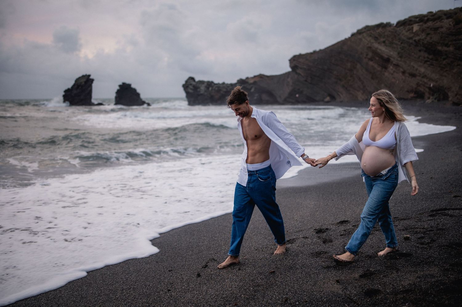 Pregnant couple walking hand in hand on black sand beach with dramatic cloudy sky and rock formations.