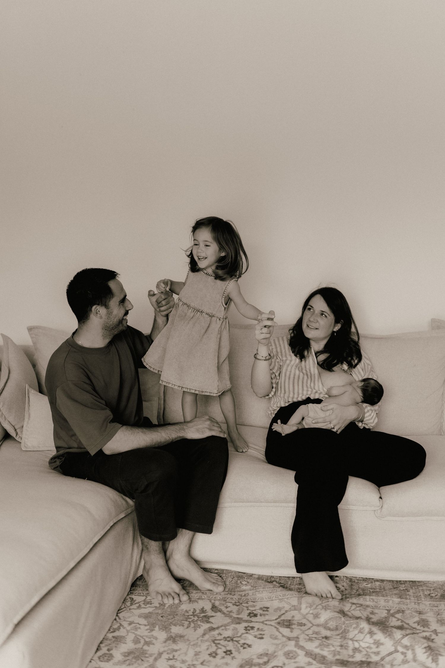 Black and white family portrait with parents and toddler on white sofa holding newborn baby.
