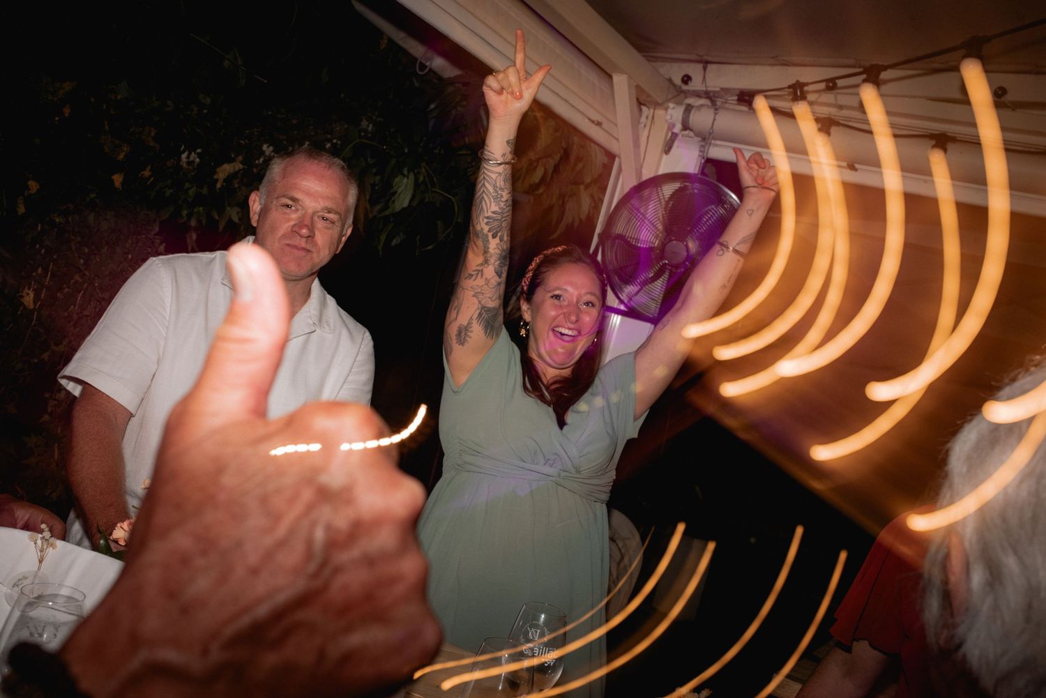Two people celebrating at a nighttime party with light trails and a thumbs up gesture in the foreground.