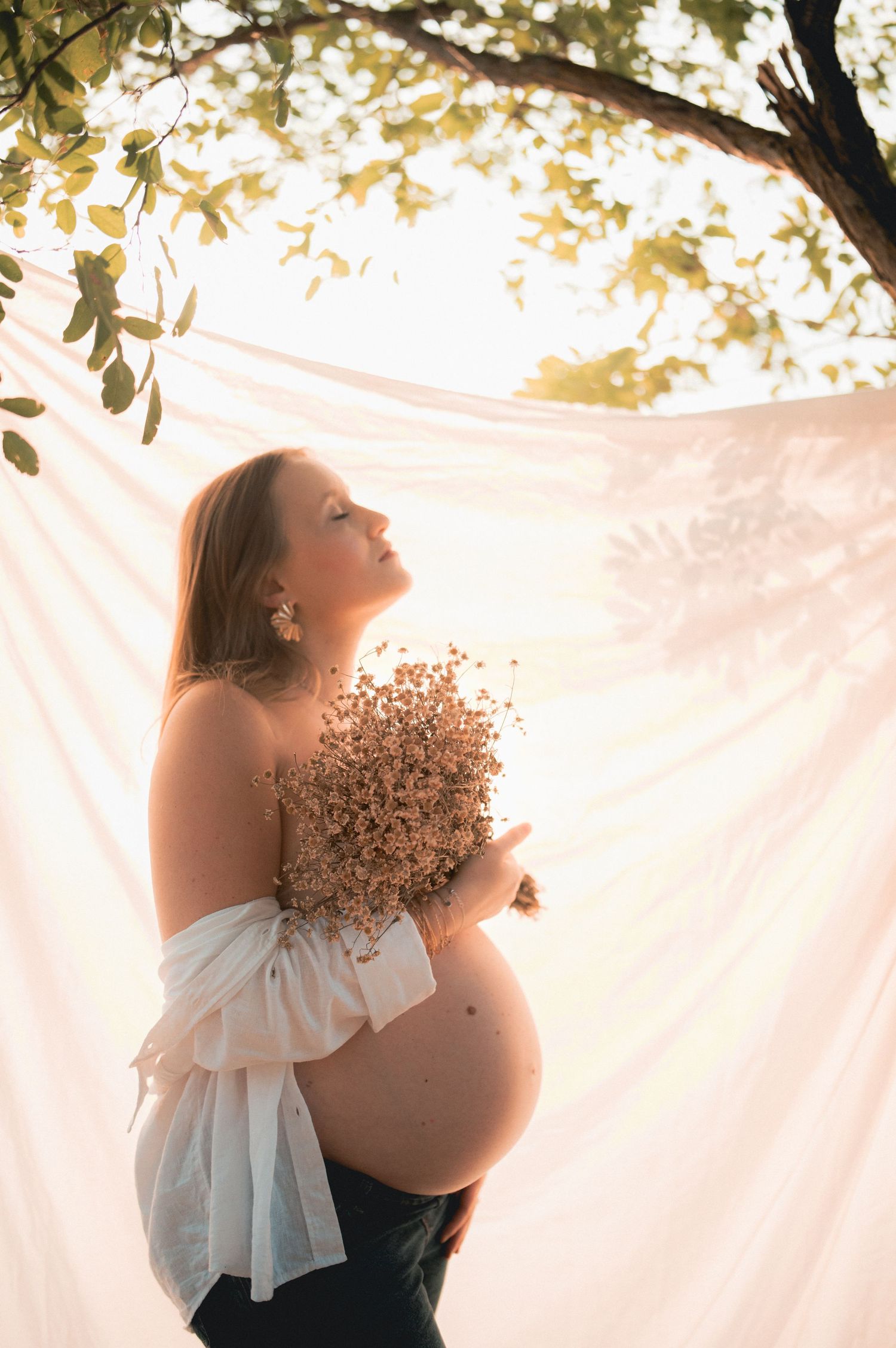 Pregnant woman holding dried flowers in golden sunlight beneath a tree with flowing white fabric backdrop.