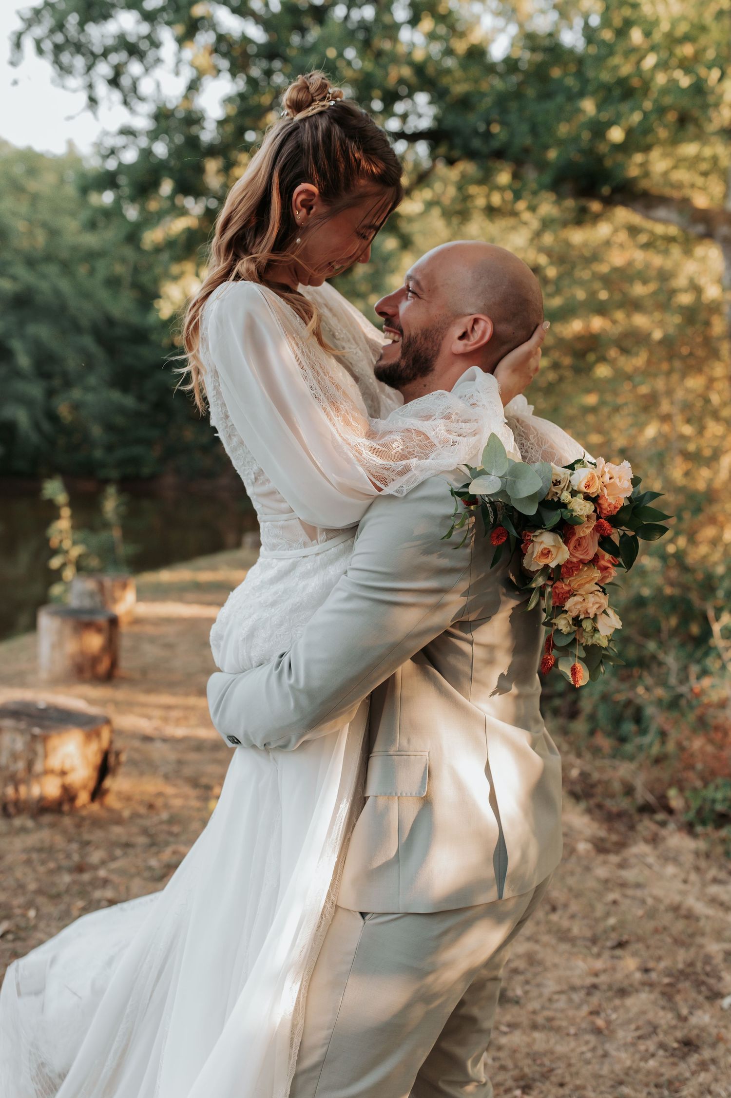 Groom in gray suit lifts bride in white lace dress, both laughing, holding bouquet, outdoors with trees.