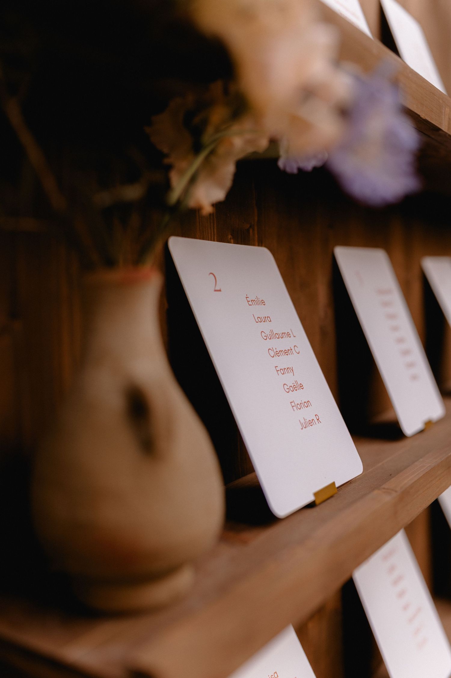 Wooden shelf displaying wedding seating chart cards with guest names and a rustic ceramic vase with dried flowers.