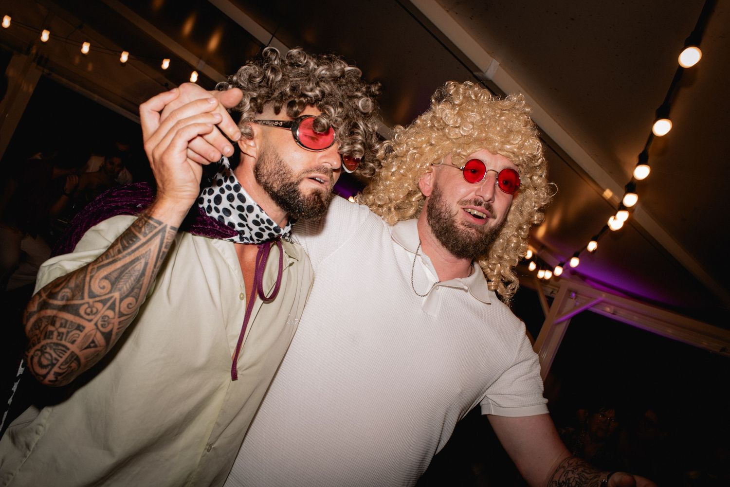 Two men in curly wigs and red sunglasses posing at a fun costume party under string lights.