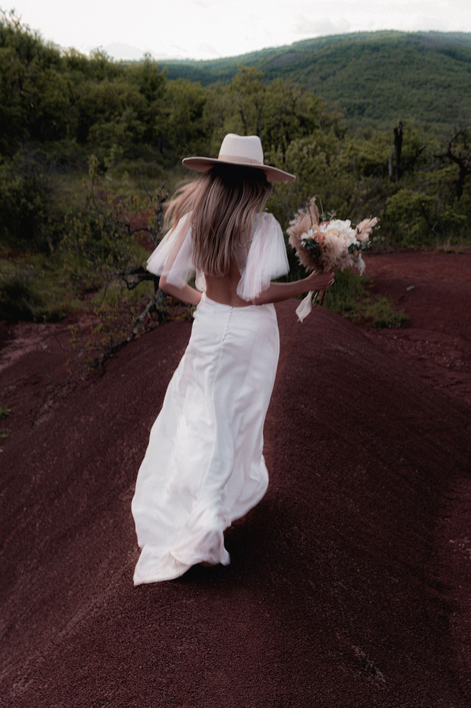 Bride in white backless gown and wide-brim hat walks red dirt path holding bouquet, lush green hills behind her.