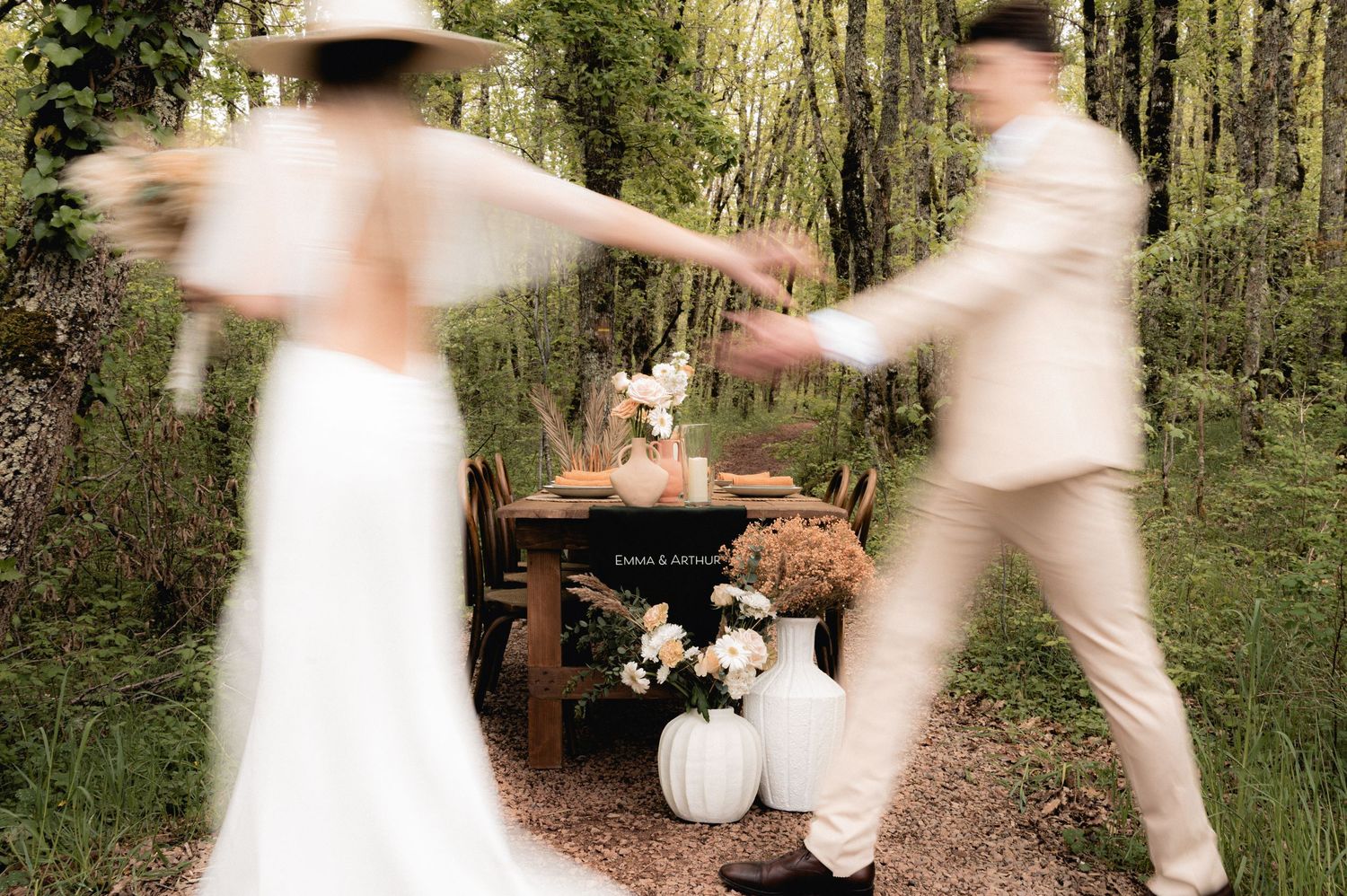 Blurred bride and groom dancing outdoors near a rustic woodland wedding table with white vases and floral decor.