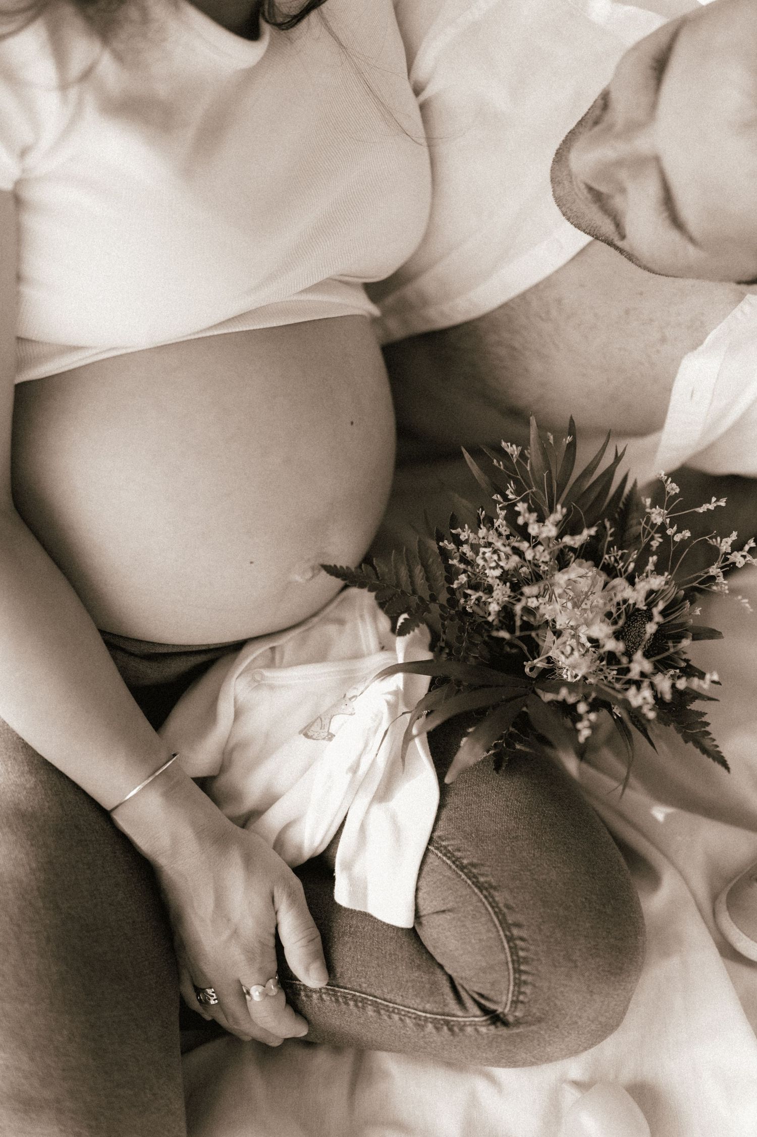 Sepia-toned maternity photo showing pregnant belly with wildflower bouquet and baby clothes.
