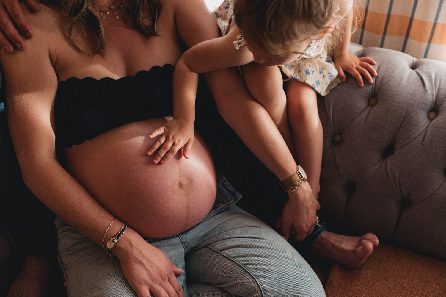 Pregnant woman in black crop top with child resting on her shoulder on a couch indoors.
