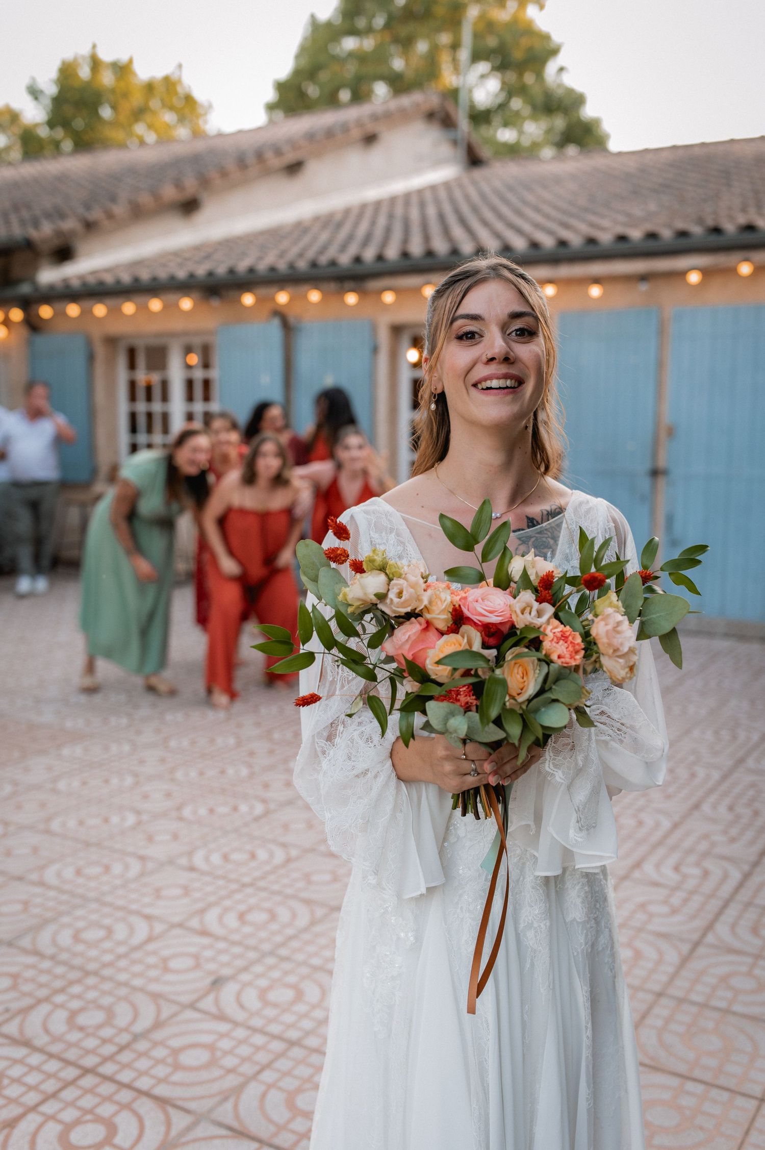 Smiling bride in white lace dress holds colorful bouquet outdoors, with wedding guests in background near rustic venue.