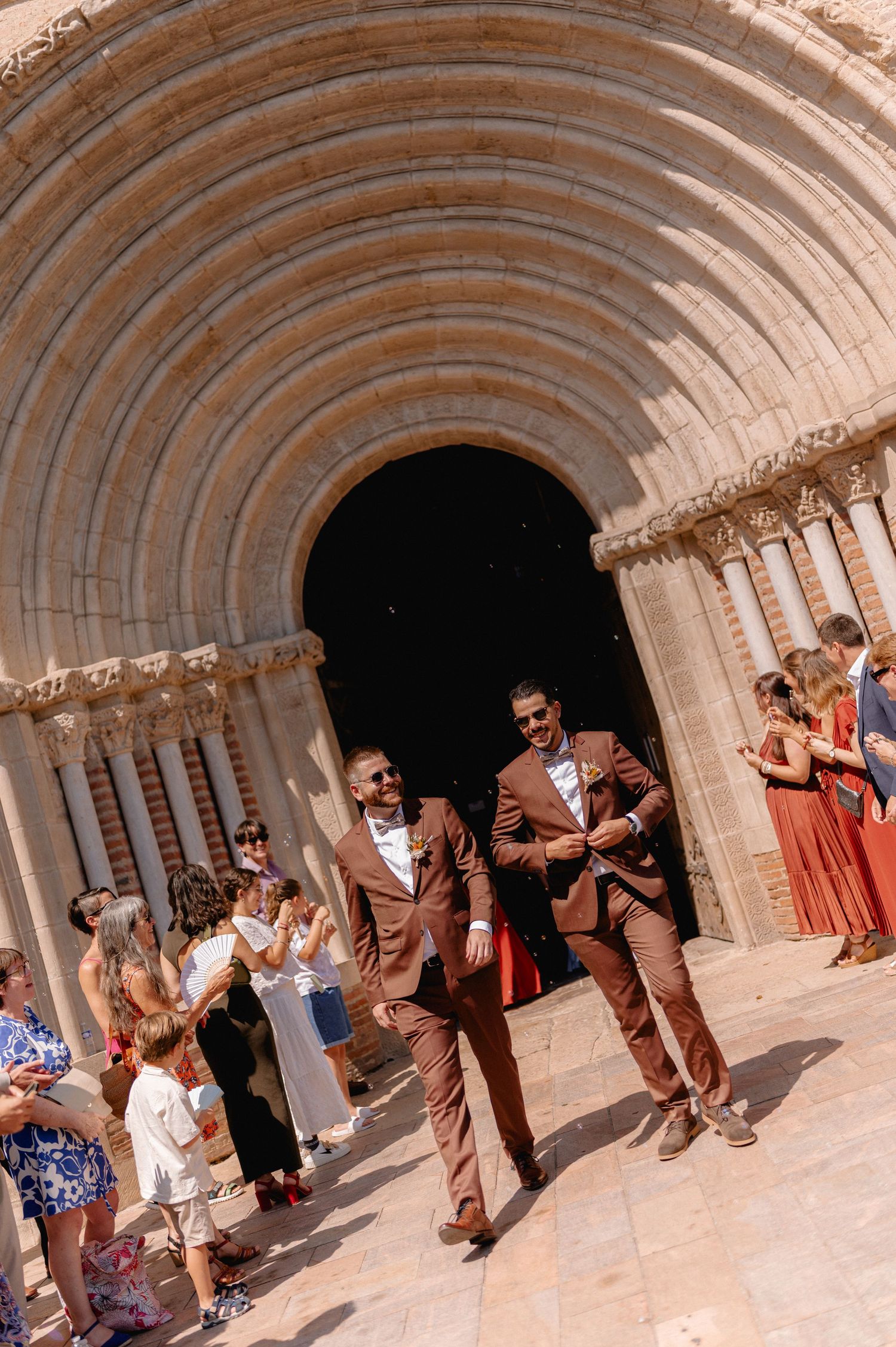 Two grooms in matching brown suits exit a stone arch church entrance, celebrated by guests on a sunny wedding day.