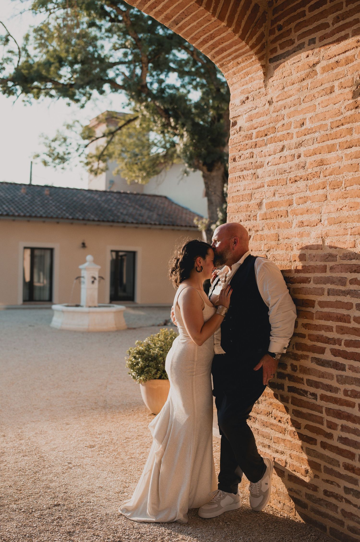 Bride and groom share a romantic kiss leaning against a rustic brick wall at a sunlit outdoor wedding venue.