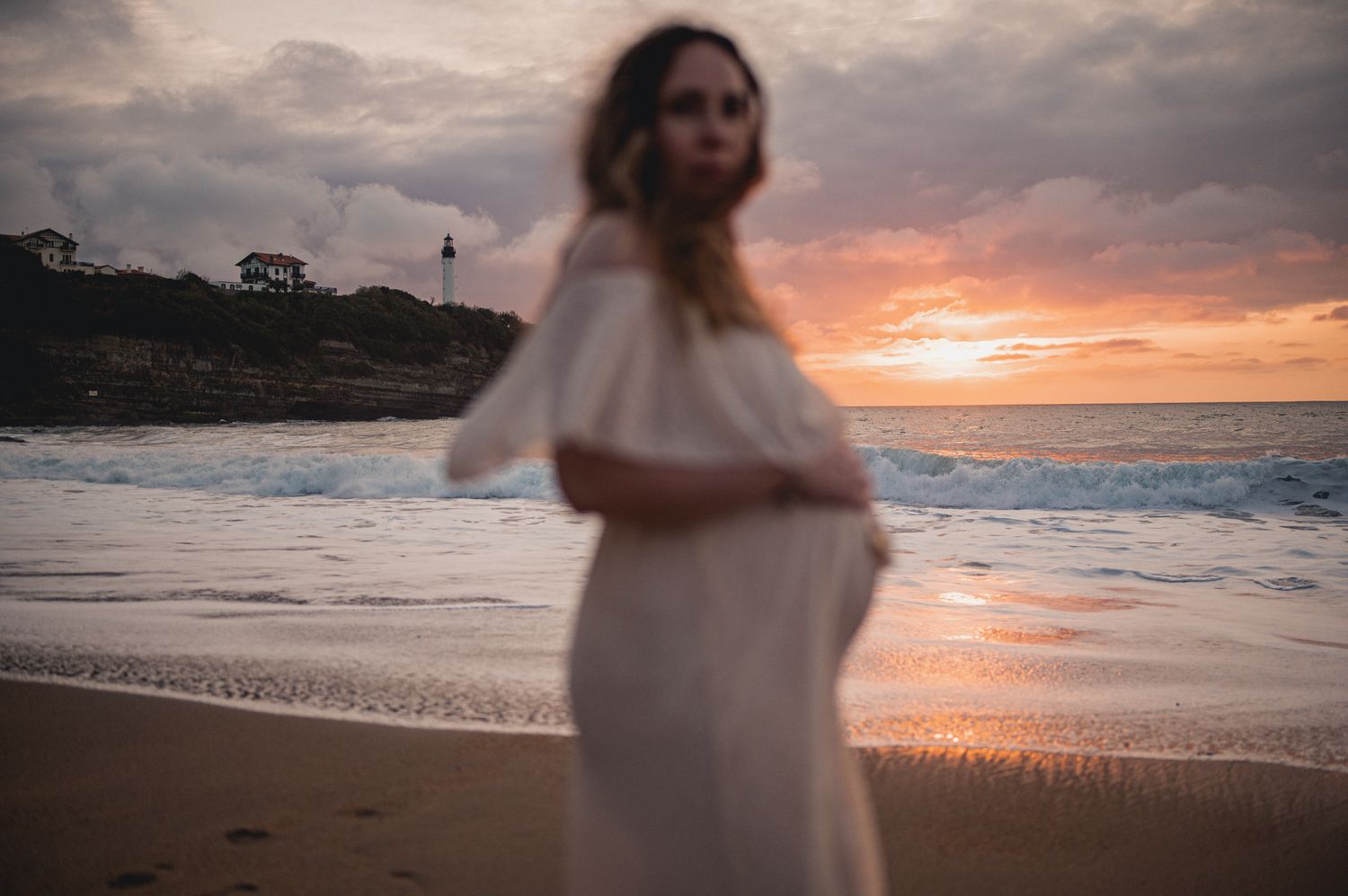 Pregnant woman in white off-shoulder dress standing on beach at golden sunset with waves behind her.