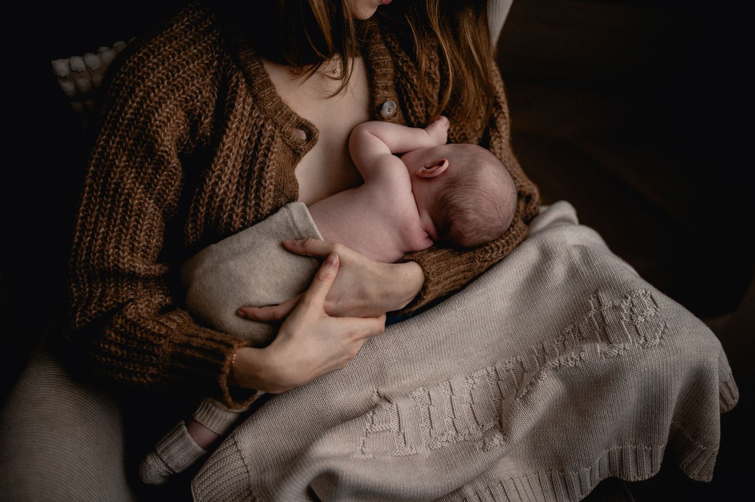 Mother in brown sweater cradling her newborn baby wrapped in a white blanket.