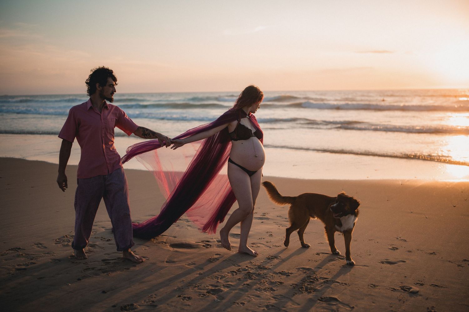 Pregnant couple walking on beach at sunset with dog, woman in flowing pink maternity gown.